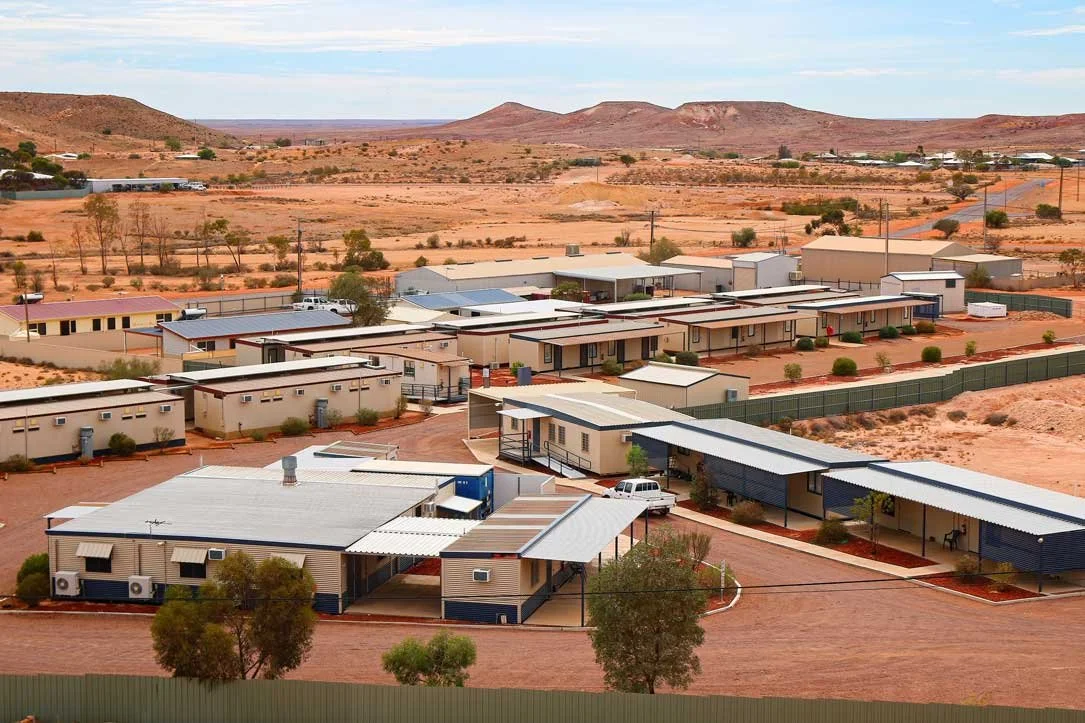 View of desert landscape with scattered bushes and hills, and a row of modern, single-story residential or community buildings with metal roofs and covered walkways.