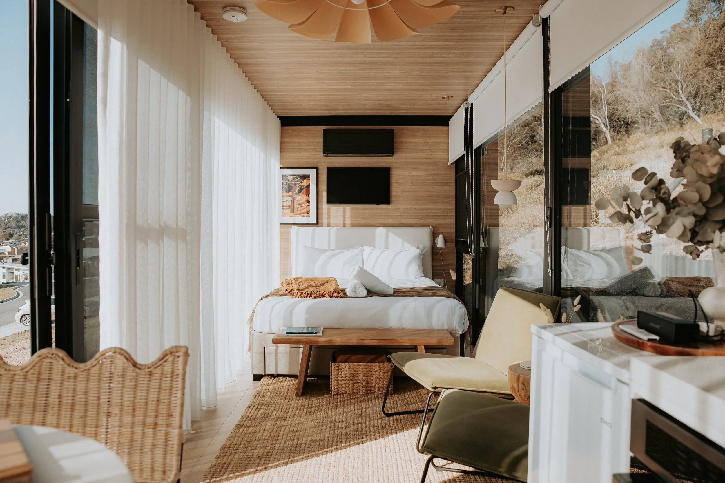 A modern bedroom with large glass windows, white curtains, a bed with white linens, a wooden headboard, a wall-mounted TV, framed artwork, a small side table, yellow chairs, and a white cabinet with decorative items.