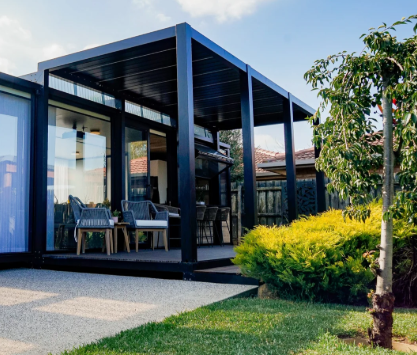 A modern backyard patio with a black pergola, outdoor furniture, and a landscaped garden with trees and bushes.