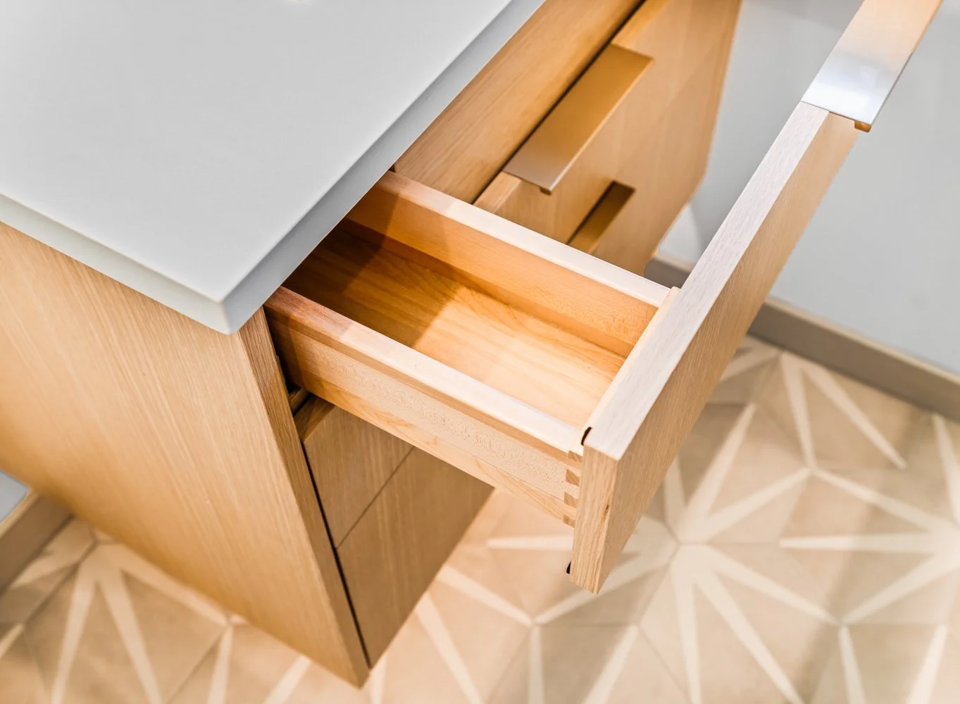 Open wooden drawer in a light-colored cabinet with a white countertop, on a patterned tile floor.