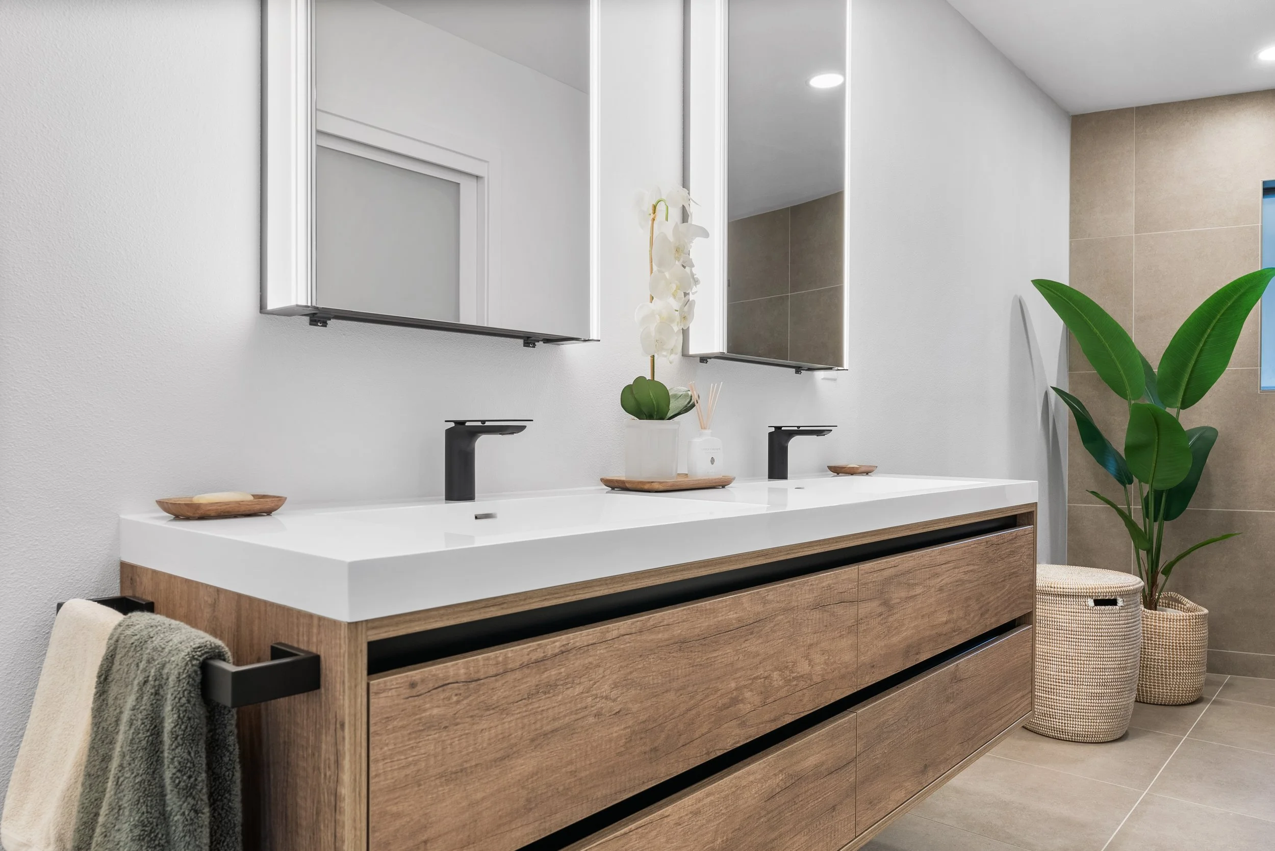 Modern bathroom with double vanity, white countertop, black faucets, large mirror, potted plant, and woven baskets.