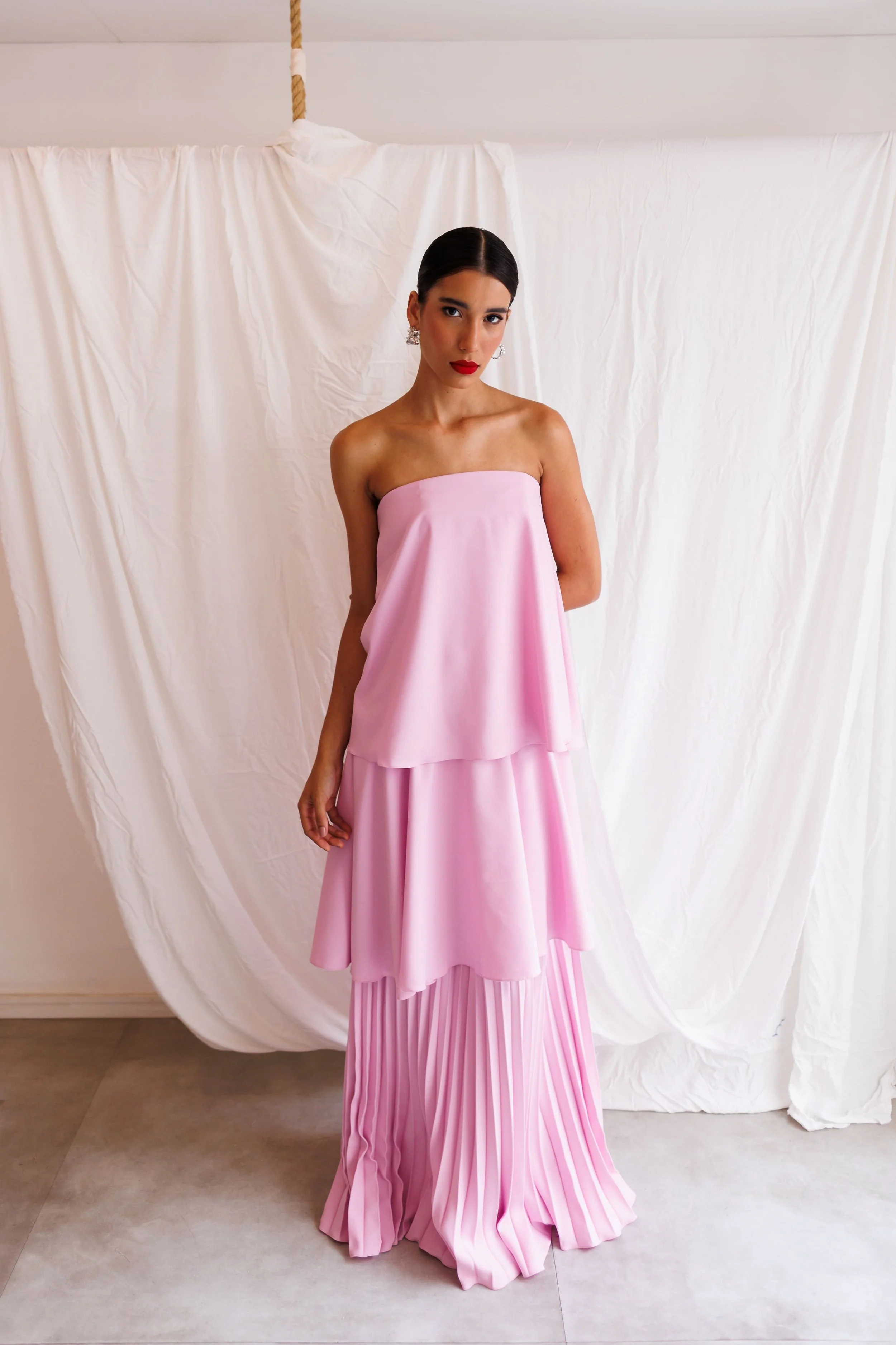 Mulher usando vestido rosa de alças finas e saia plissada, de cabelo escuro preso, usando batom vermelho, em frente a cortina branca.