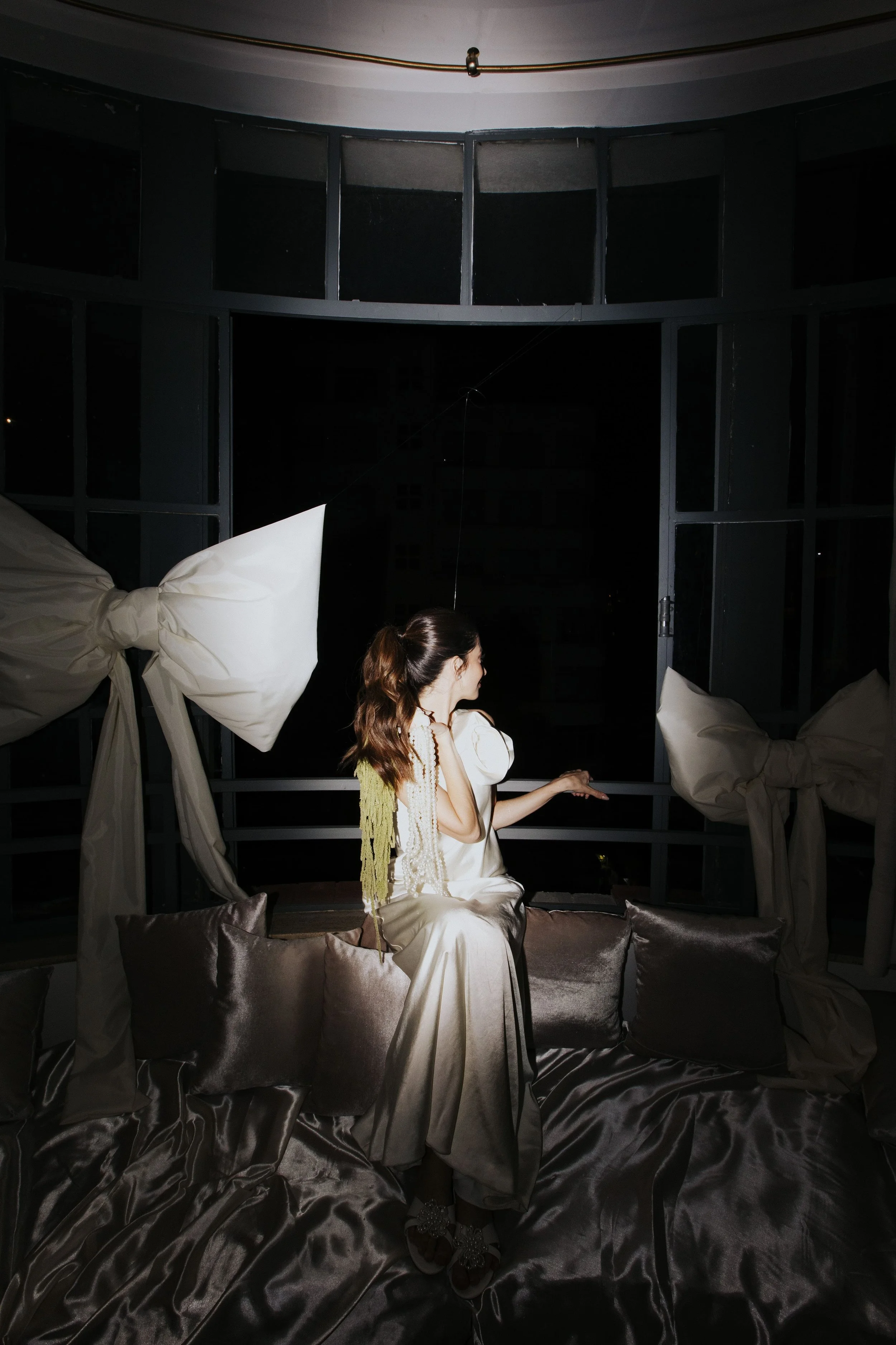 Mulher de cabelos castanhos claros amarrados, vestida com vestido de cetim branco, sentada em um sofá de veludo cinza, em um ambiente com janelas e cortinas em formato de laço, durante a noite.