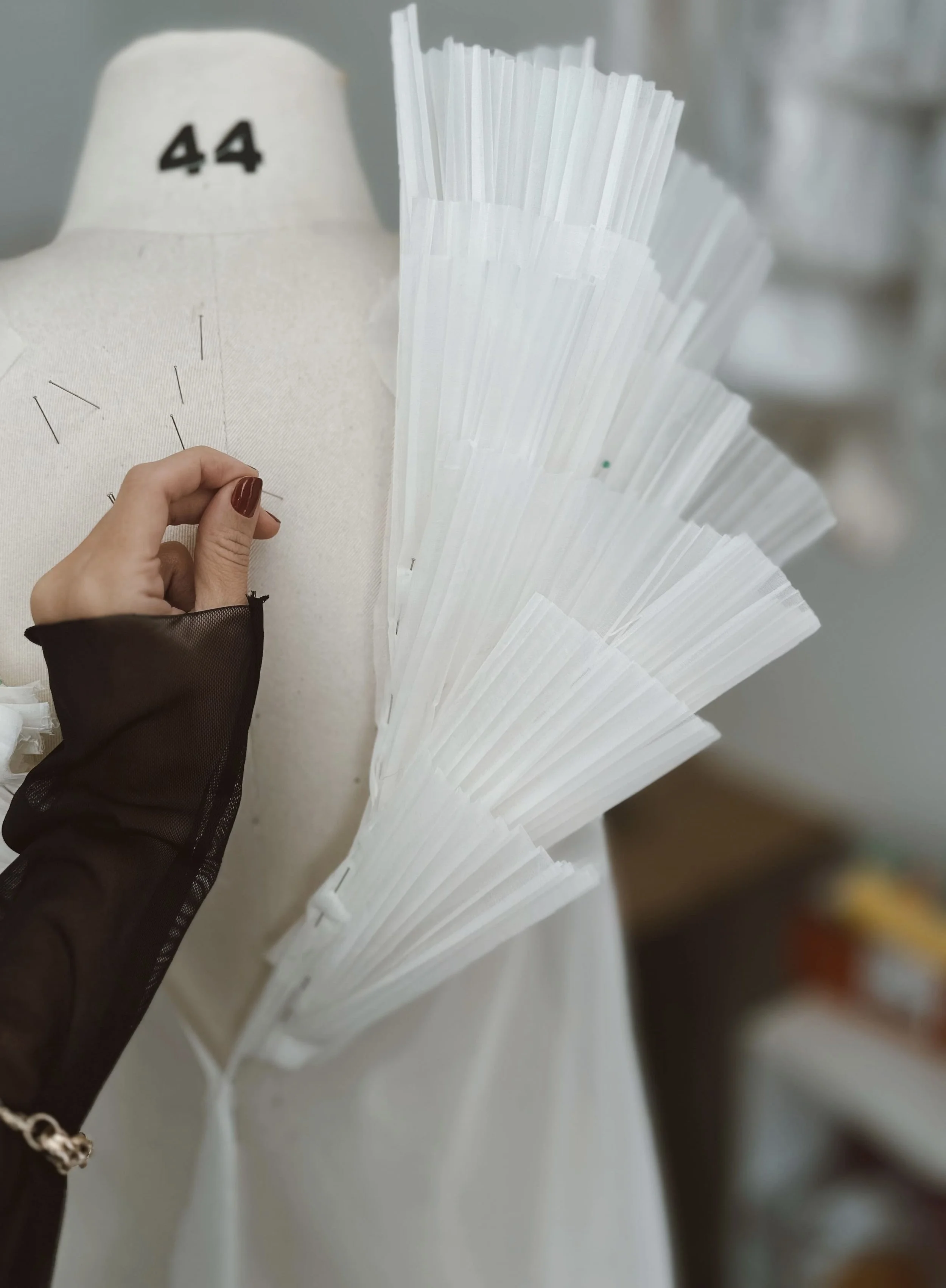 Certo, aqui está a descrição em português: Uma manequim de costura com a numeração 44, com pedaços de tecido ou papel branco em forma de leque presos na lateral do manequim, enquanto uma pessoa com unhas pintadas de marrom está ajustando uma agulha ou alfinete na área do busto do manequim.