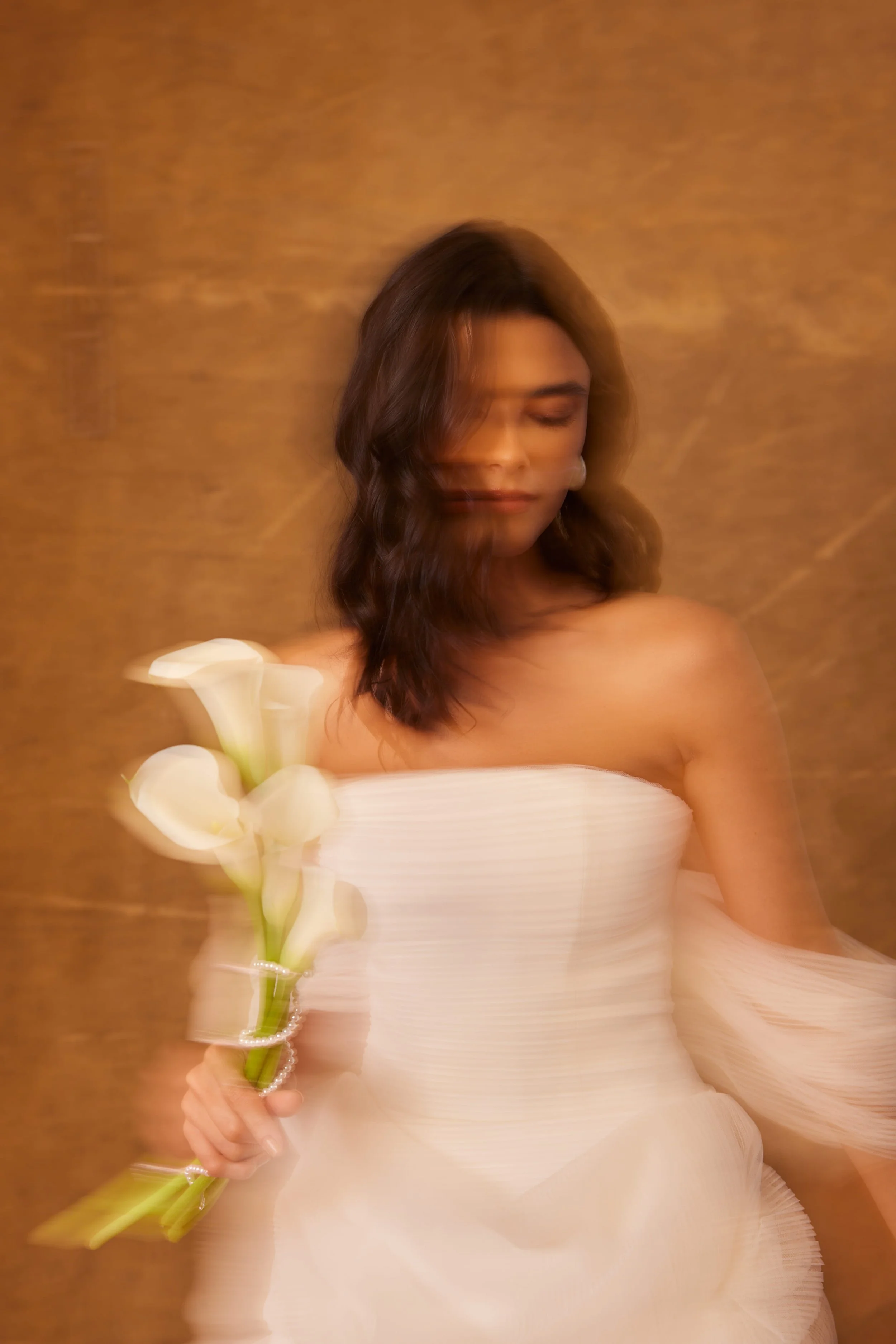 Mulher com vestido branco e cabelo castanho escuro, segurando um ramo de flores brancas, com efeito de movimento na fotografia, fundo dourado.