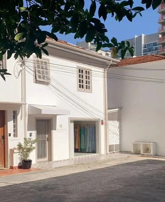 Edifício branco com uma loja de roupas na rua, árvores na frente, céu claro e edifícios altos ao fundo.