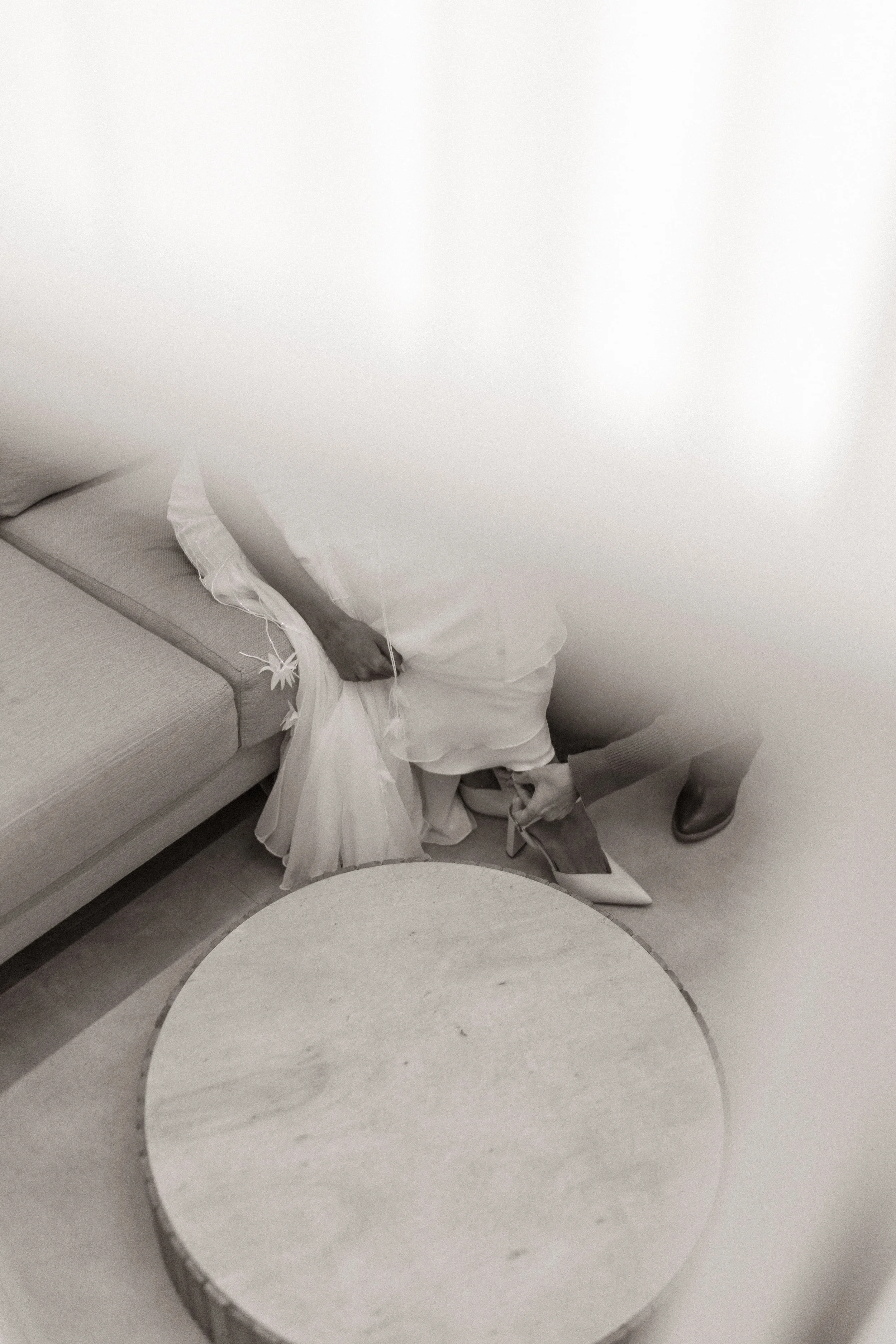 Pessoa ajustando o salto de um sapato de salto fino, sentado em uma cadeira ao lado de um sofá, em um ambiente interno.