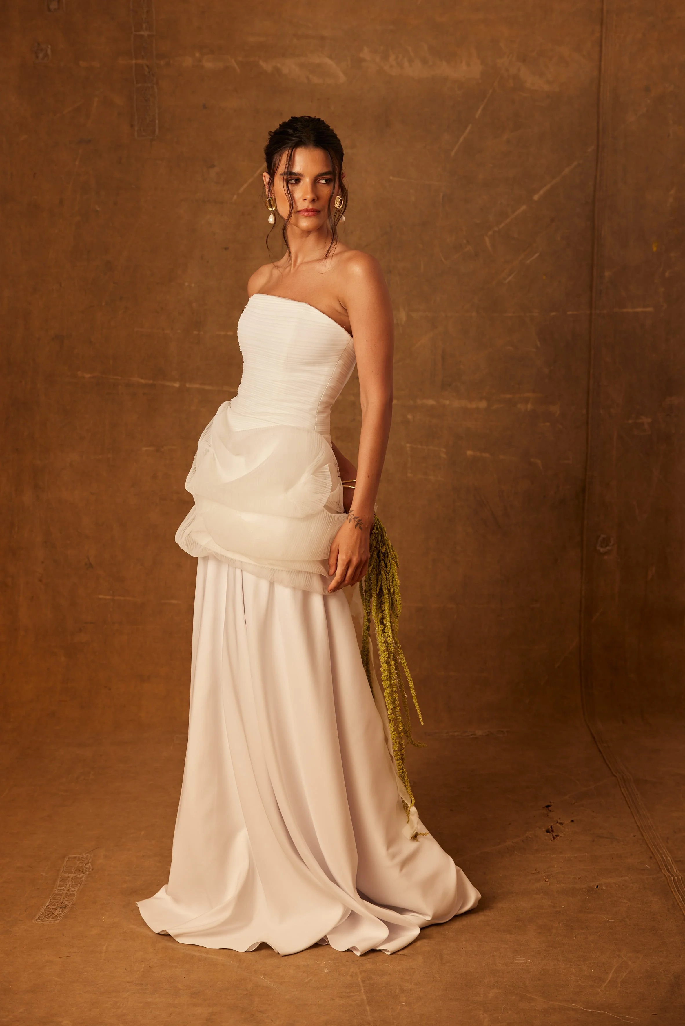 Mulher vestida com vestido de festa branco, com saia longa e detalhes em tecido emplumado, com acessórios dourados e brincos grandes, posando em fundo marrom.