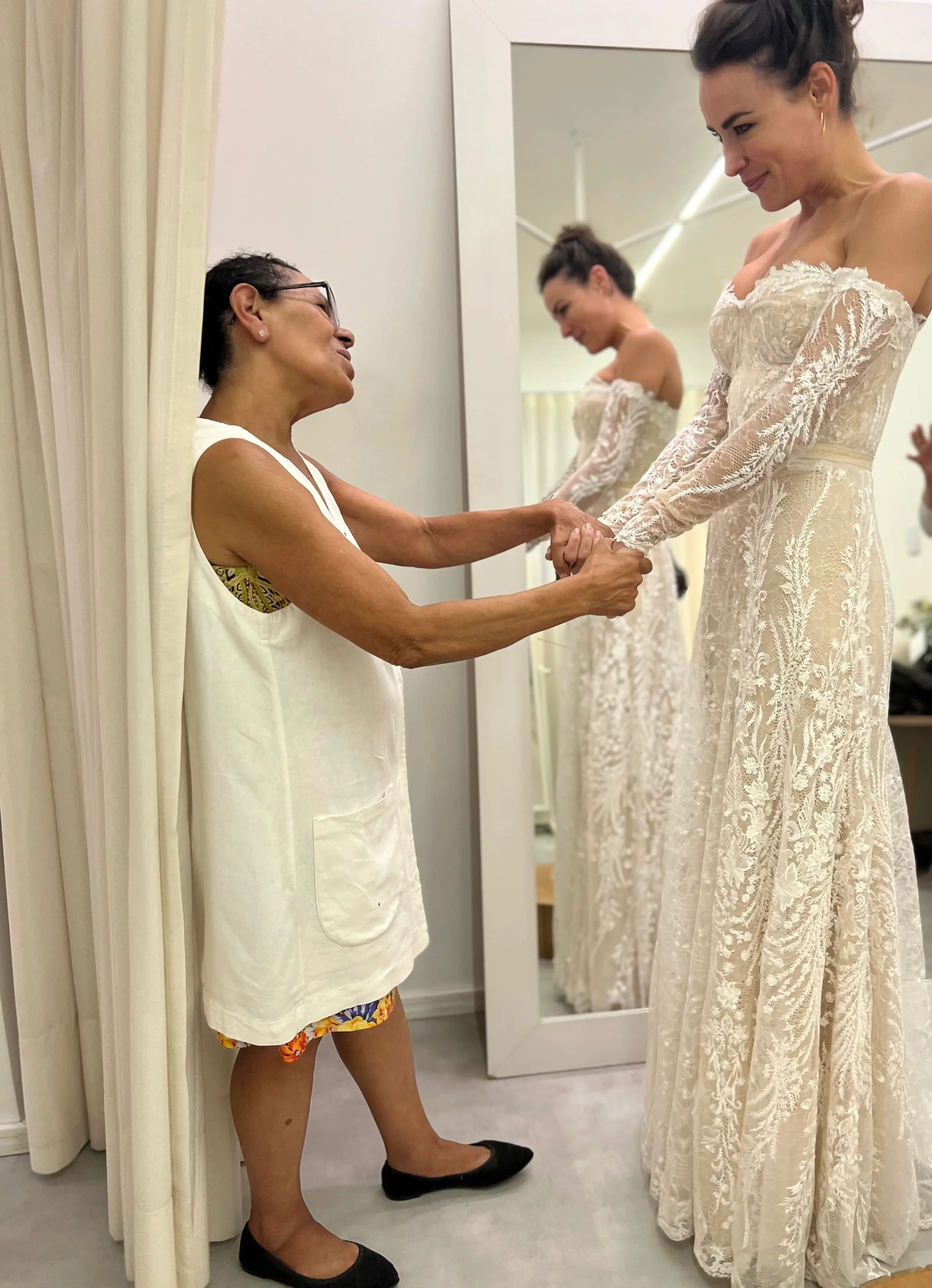 Mulher mais velha ajudando uma mulher jovem a experimentar um vestido de casamento em uma loja. Elas estão felizes, segurando as mãos e sorrindo.