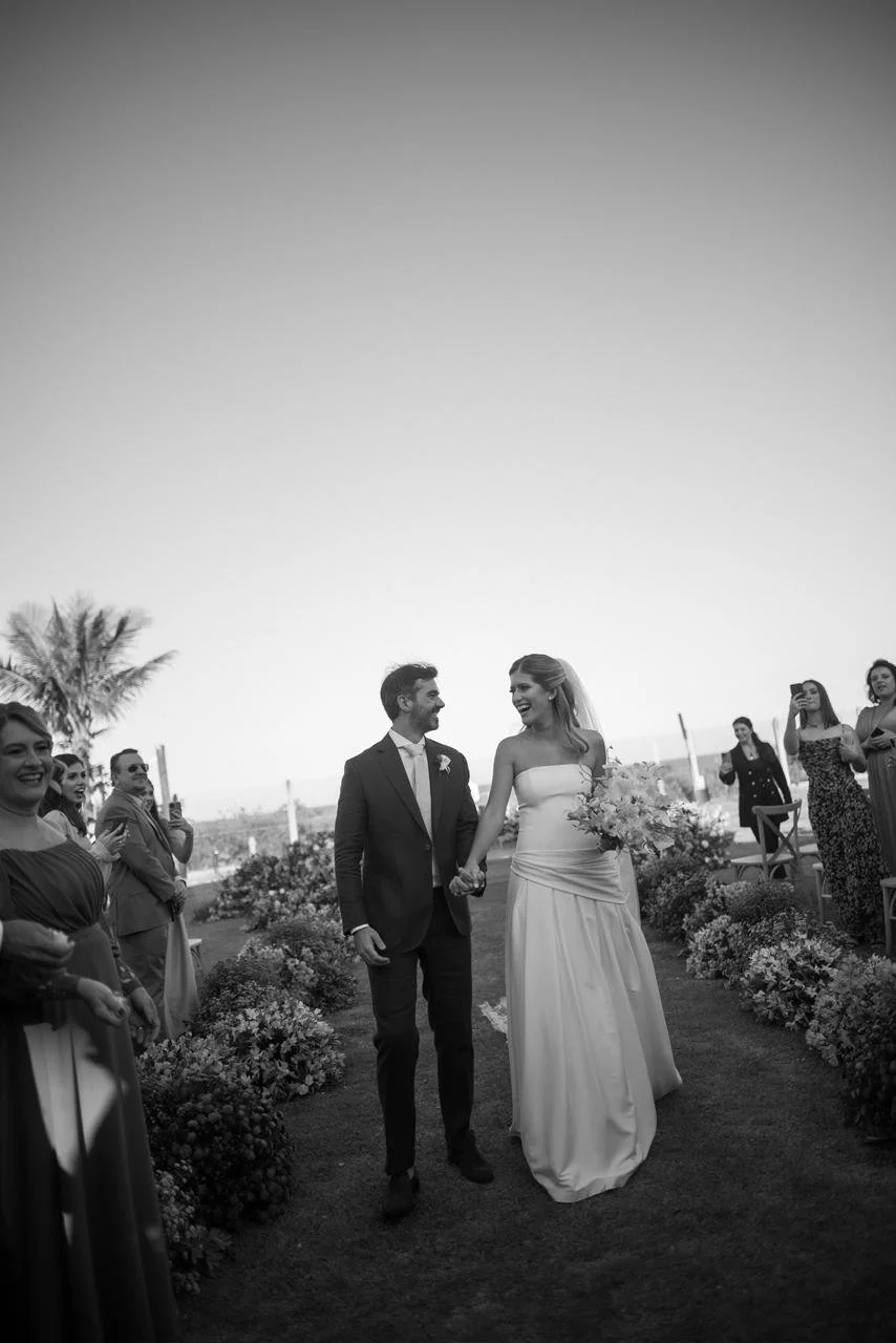 Casal de noivos no casamento ao ar livre, segurando as mãos, cercados por convidados, flores e plantas, sob um céu claro