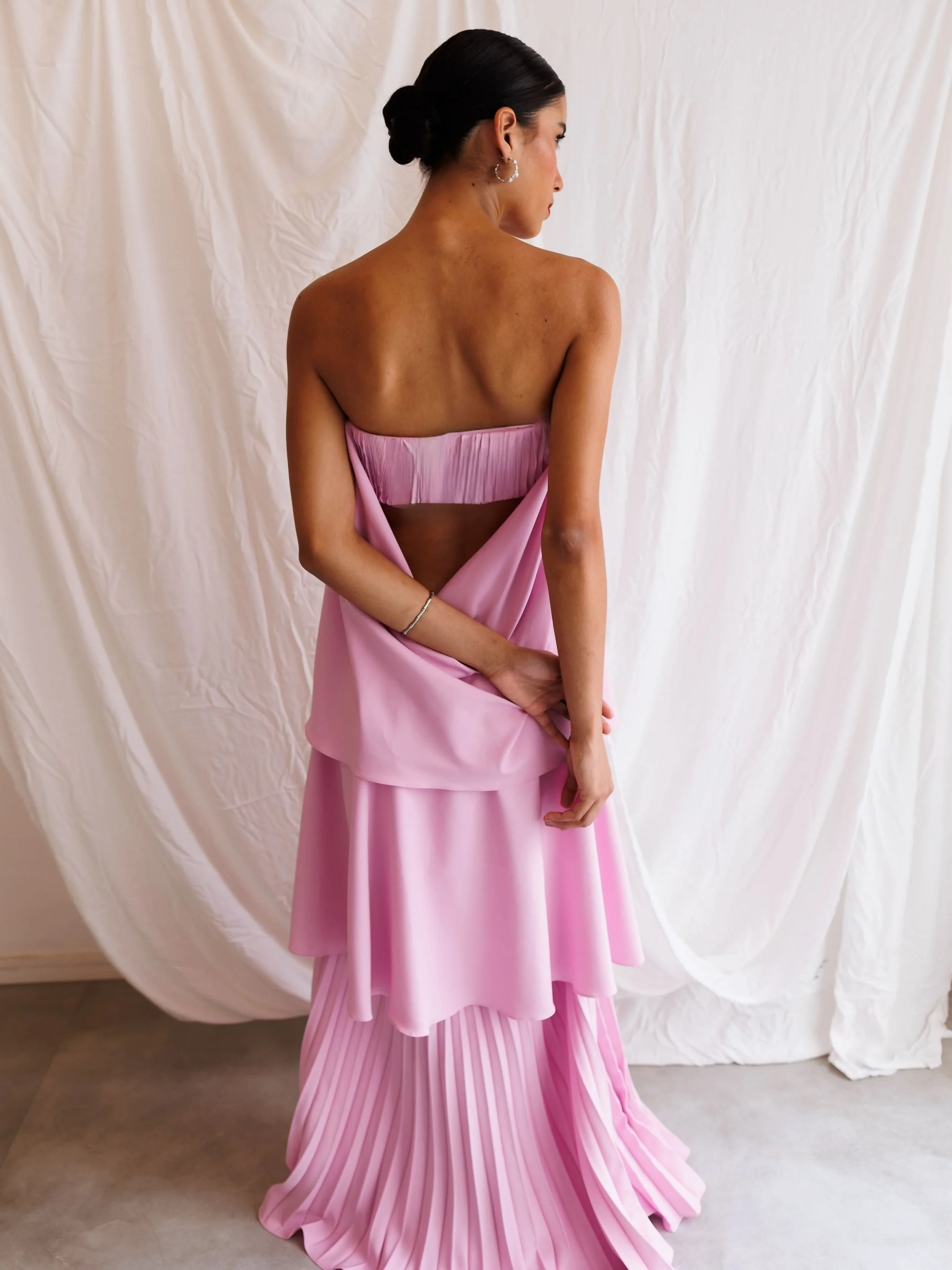 Mulher com pele morena, cabelos escuros presos em coque, usando brincos de argola prateados, vestindo saia rosa claro com parte superior de tecido acetinado e saia plissada, posando de costas com uma cortina branca ao fundo.