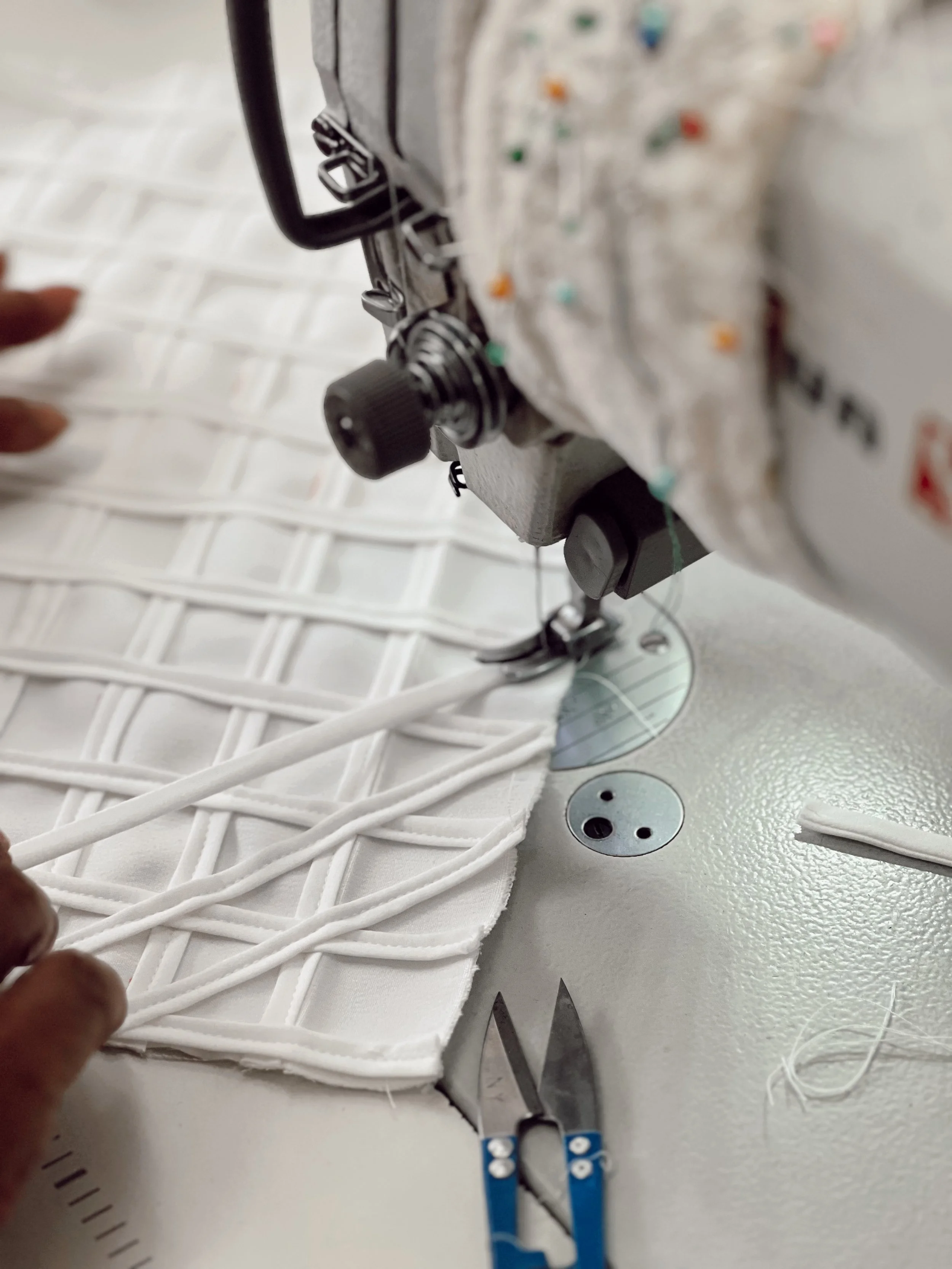 Detalhe de uma máquina de costura industrial costurando tecido branco, com mãos auxiliando e um par de tesouras na mesa.