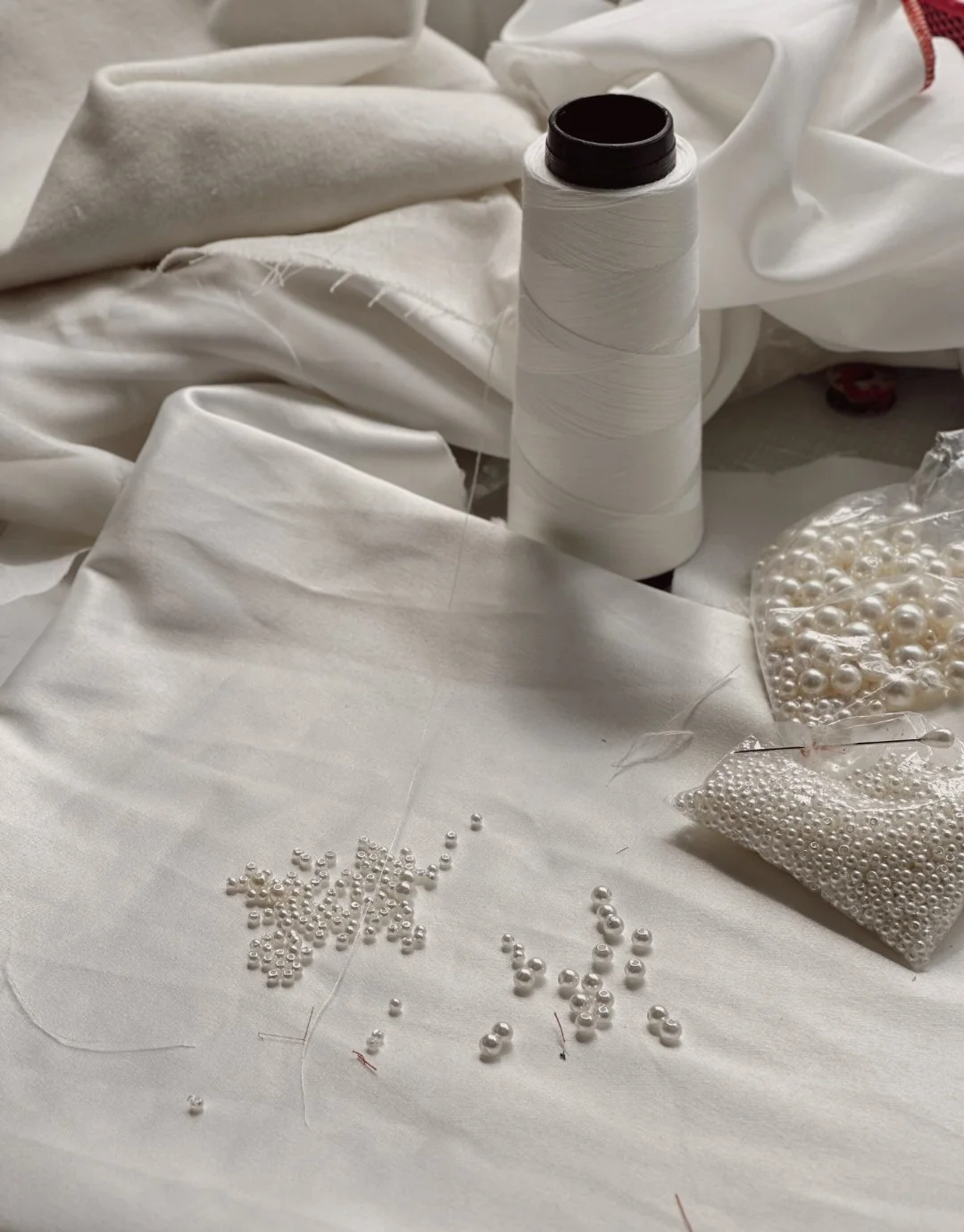 Tecidos brancos, uma bobina de linha branca, pérolas brancas e ferramentas de costura em uma superfície.