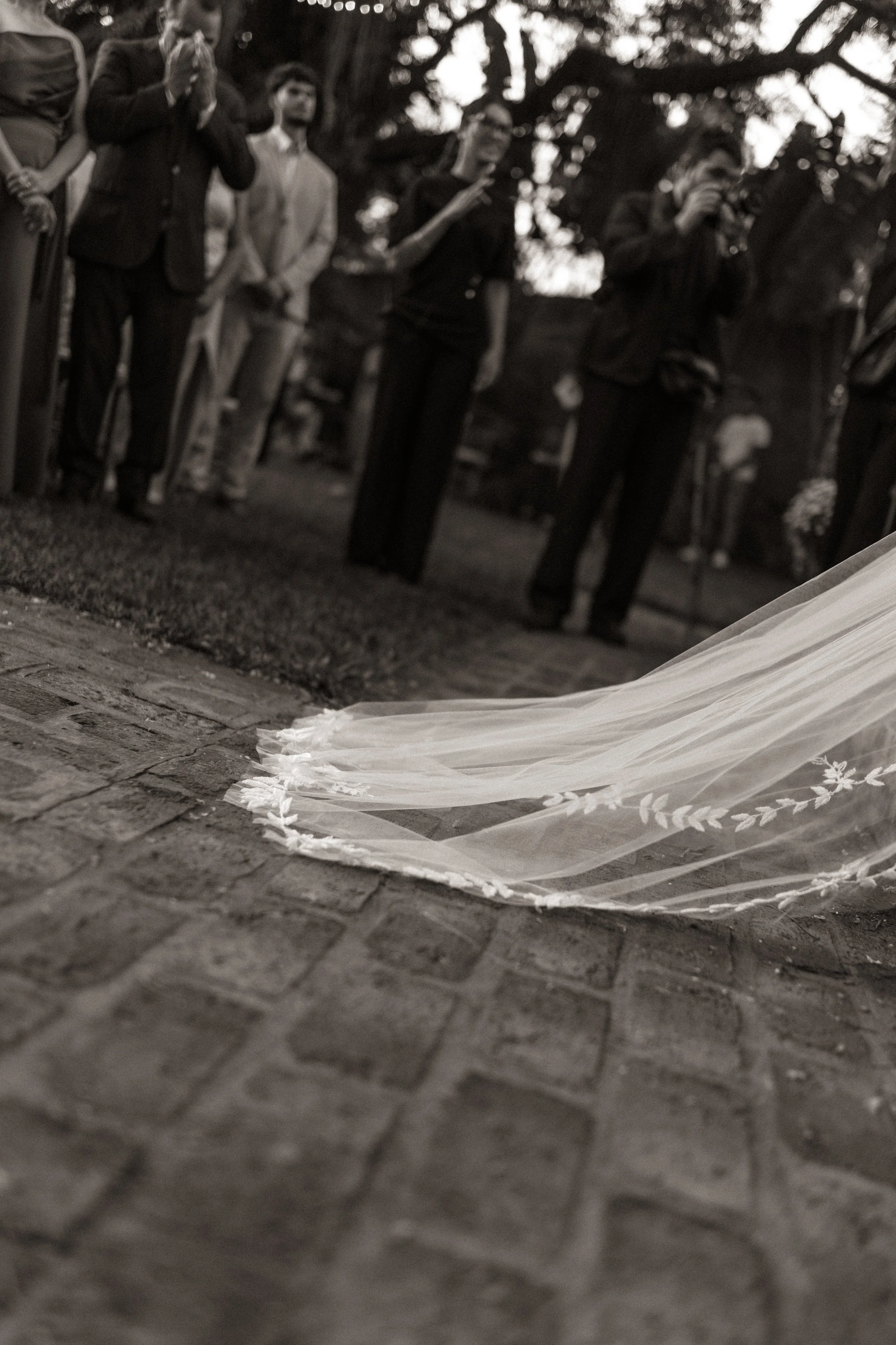Simulação de vestido de noiva no chão diante de convidados em um casamento ao ar livre, em preto e branco.