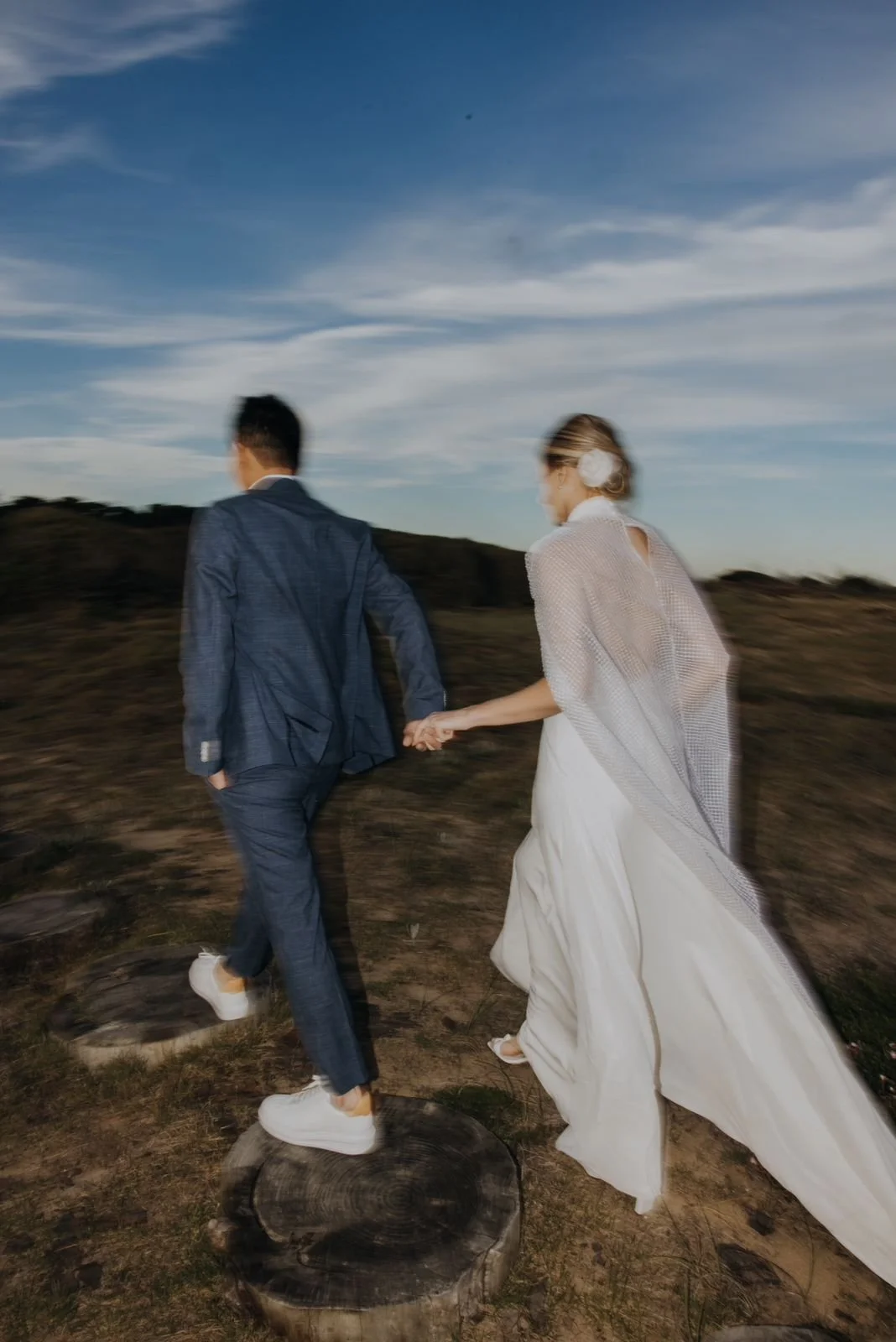 Casal de casamento andando de mãos dadas em campo ao pôr do sol, com céu azul e nuvens finas.