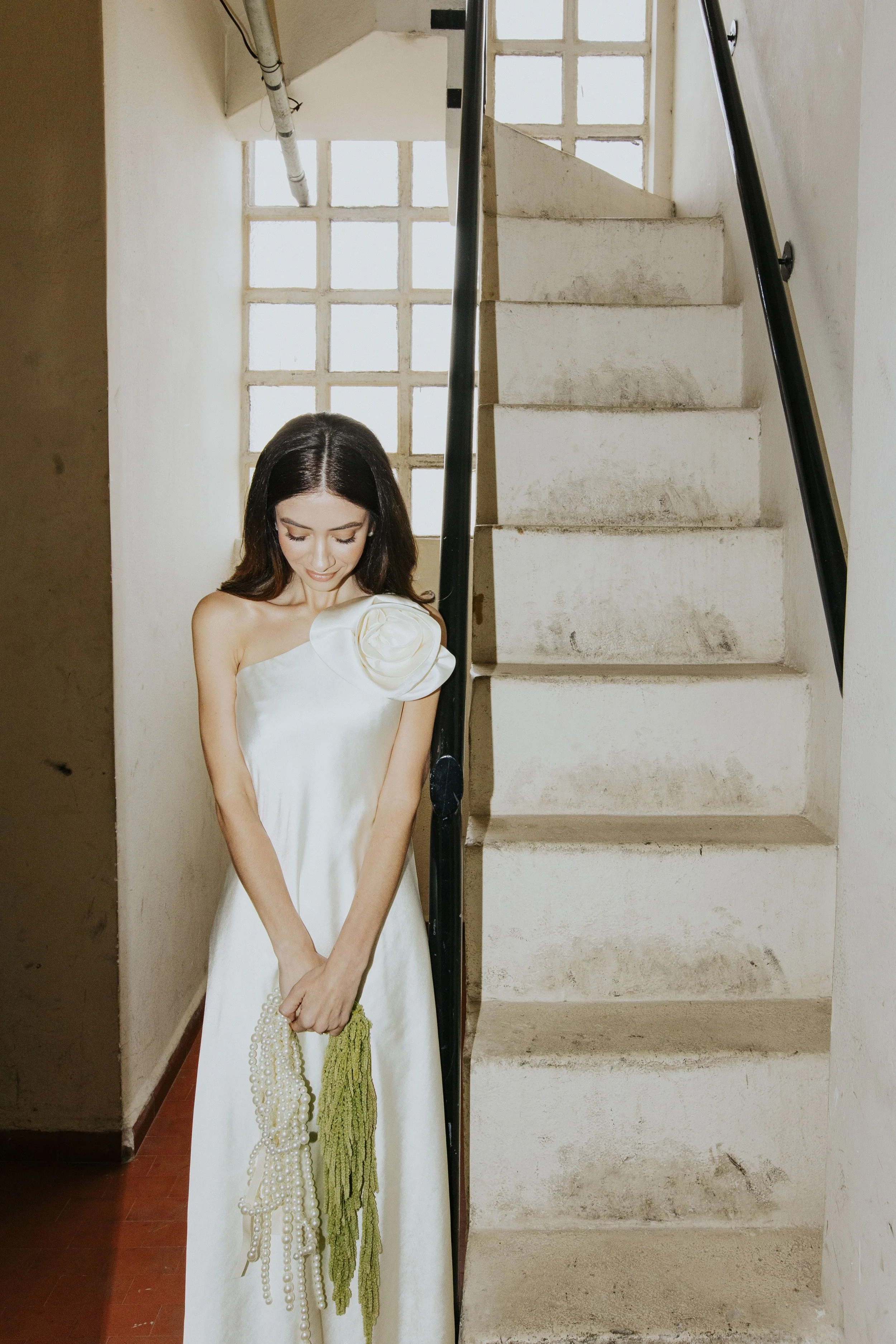 Mulher com vestido branco, de cabelo escuro, segurando bolsas de pé ao lado de escadaria com parede de blocos de vidro ao fundo.