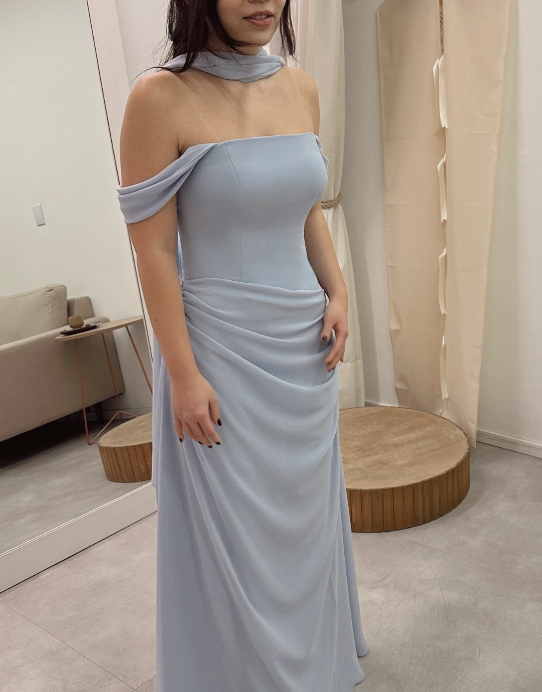 Mulher usando vestido de cerimônia azul claro, com detalhes drapeados e ombros descobertos, em um ambiente interno.