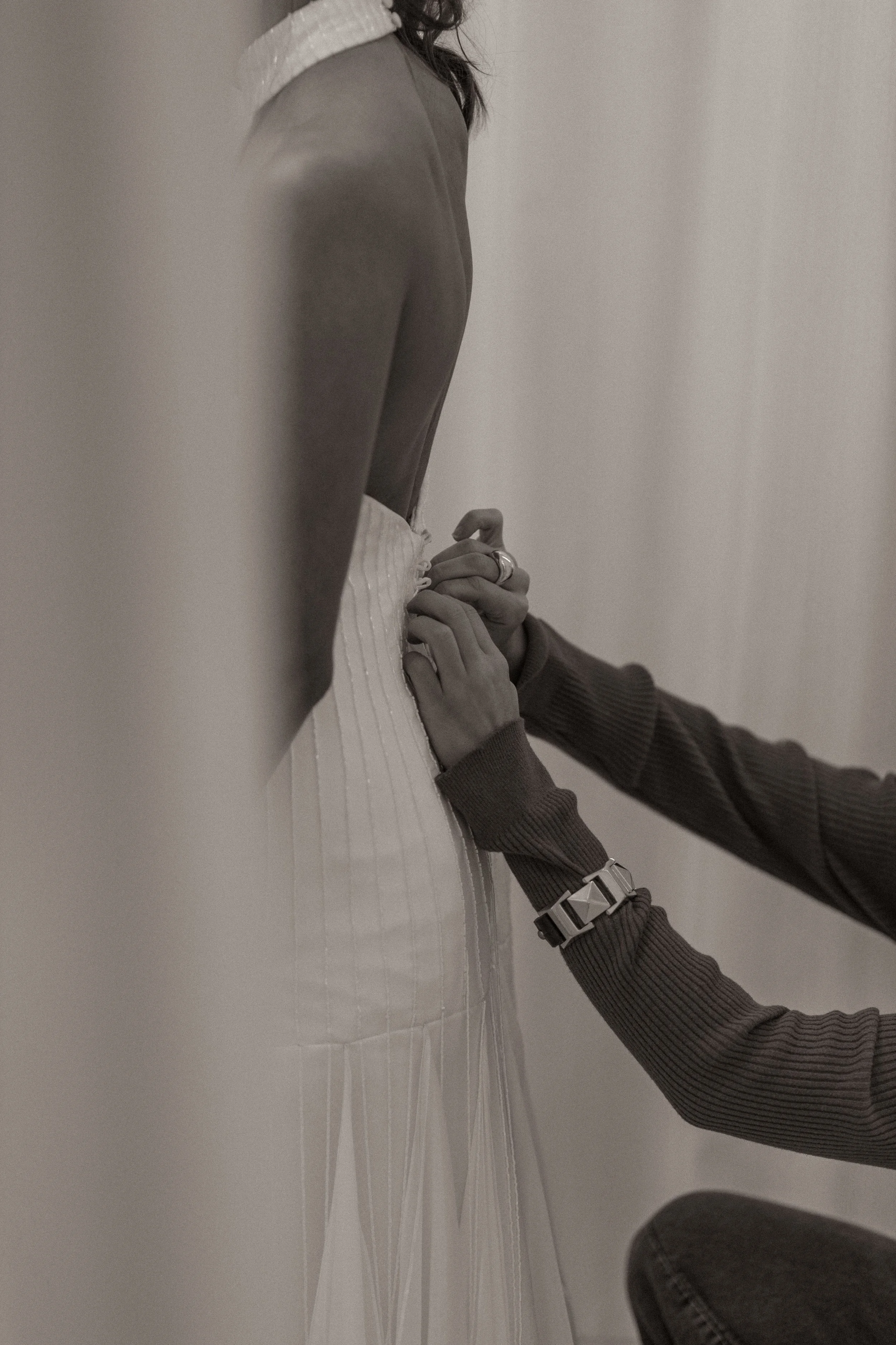 Pessoa ajustando vestido de noiva com mãos usando pulseira e anéis, imagem em tons de cinza.