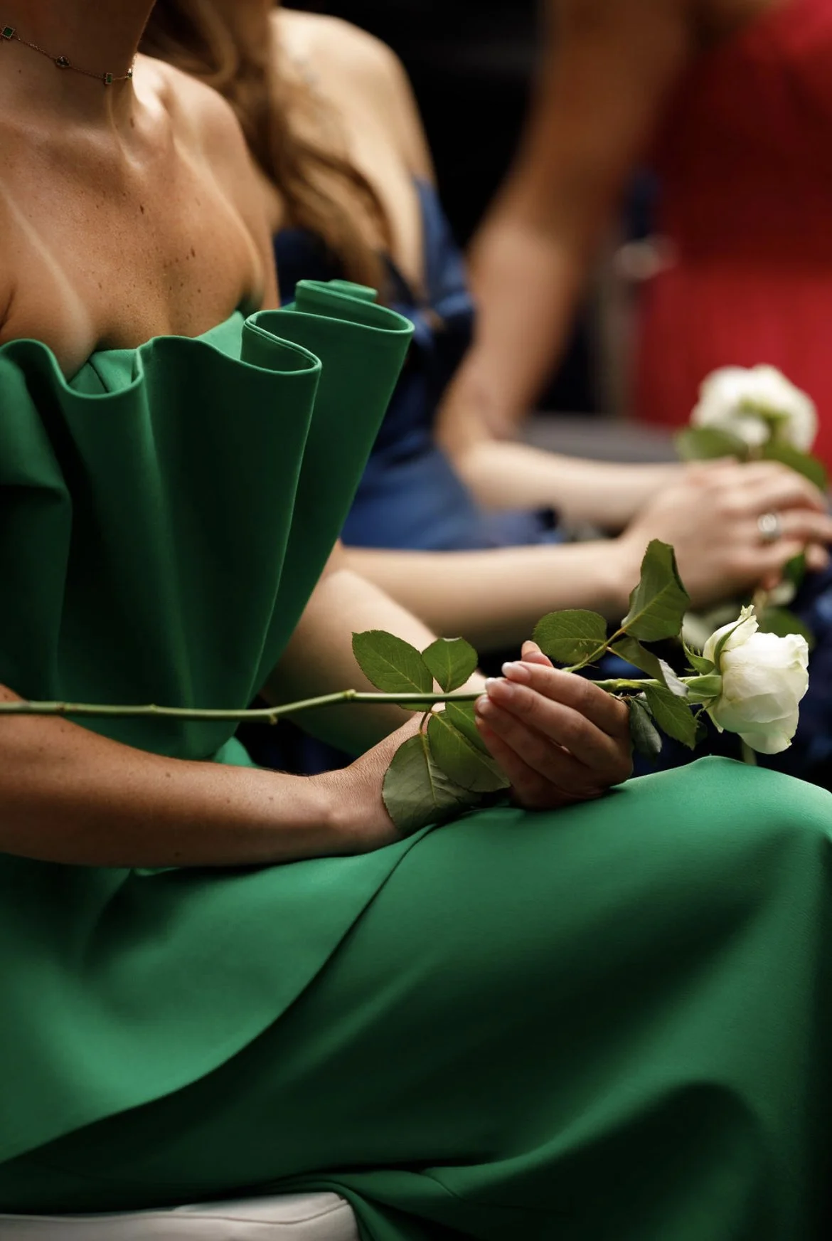 Mulher vestida com vestido verde vibrante, segurando uma rosa branca com folhas verdes. Outras pessoas ao fundo, também com roupas elegantes e segurando rosas, em um ambiente possivelmente formal ou de evento.