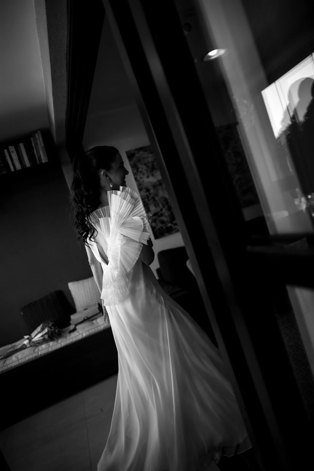 Mulher com vestido de noiva e acessórios, segurando um leque, observando para fora de uma janela dentro de um ambiente interno.