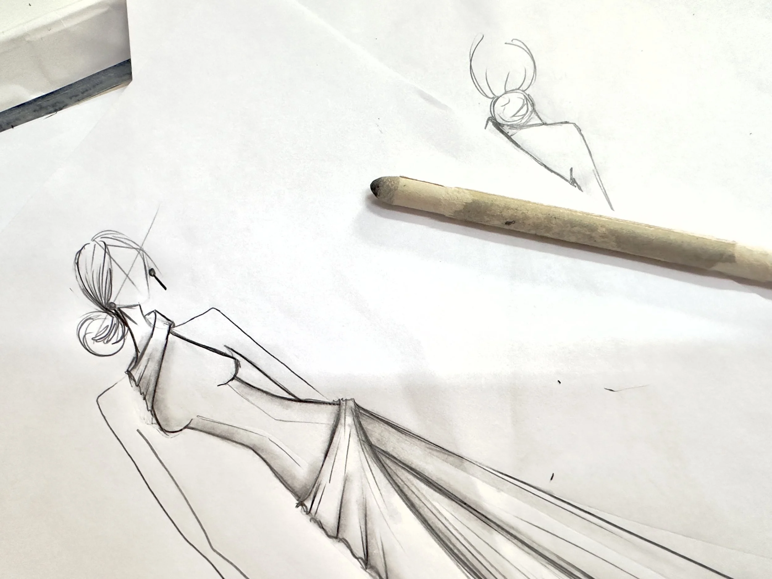 Esboço de uma figura feminina de chapéu, com cabelo preso em um coque, vestindo um vestido longo, feito com lápis sobre papel branco, com um pedaço de carvão ou carvão vegetal ao lado.