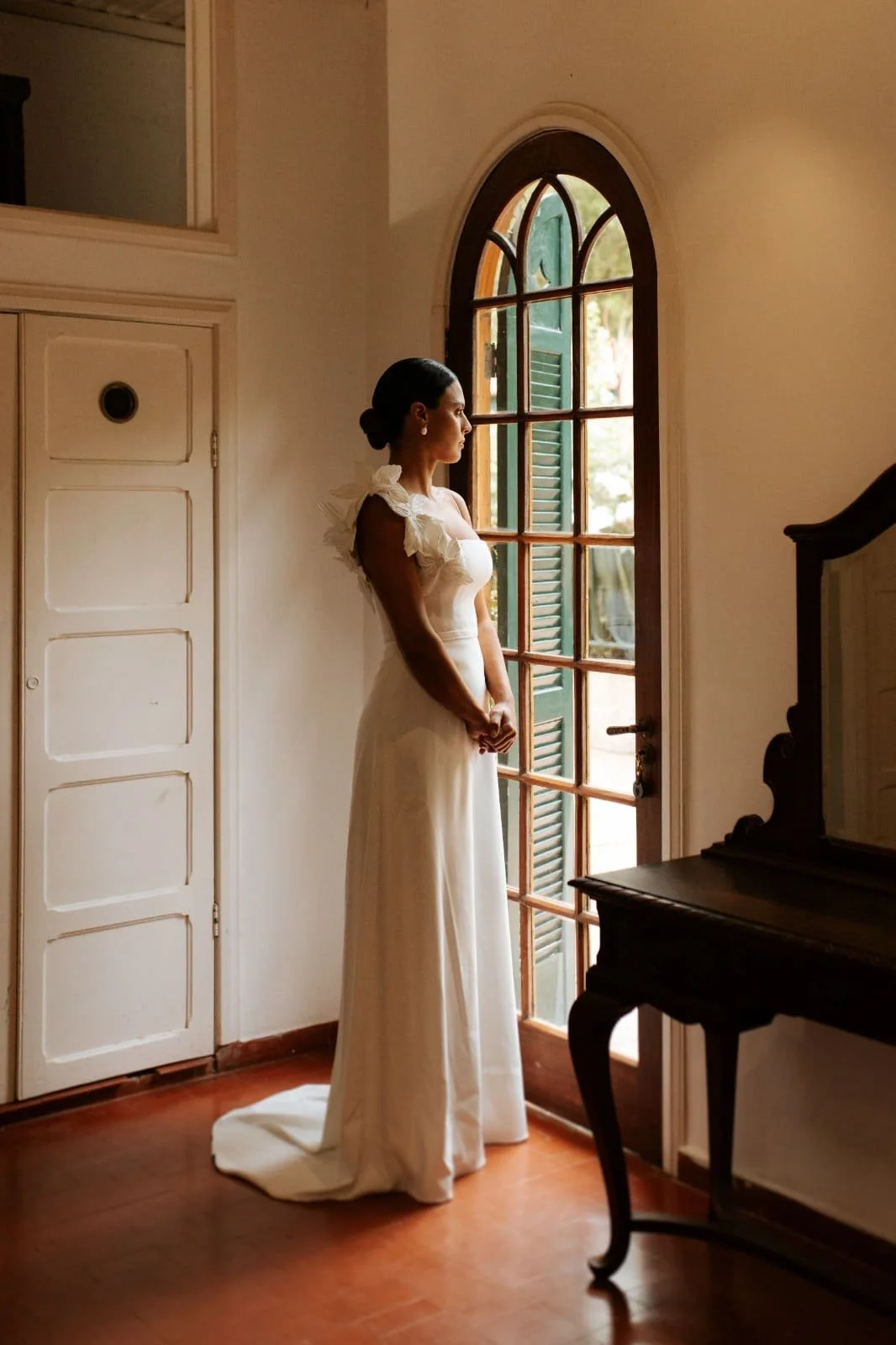 Mulher vestida de branco em um vestido de noiva, de cabelo preso, olhando para fora por uma janela de madeira com vidro, dentro de uma sala com piso de madeira e móveis clássicos.