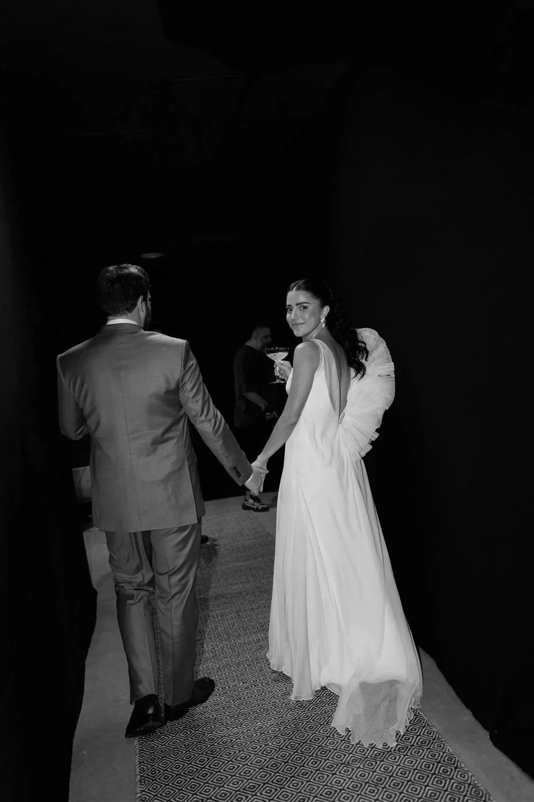 No corredor de um evento, um casal de noivos de mãos dadas, um homem de terno cinza e uma mulher de vestido branco, ambos de costas, olhando para a câmera, enquanto uma pessoa ao fundo segura uma taça de coquetel.