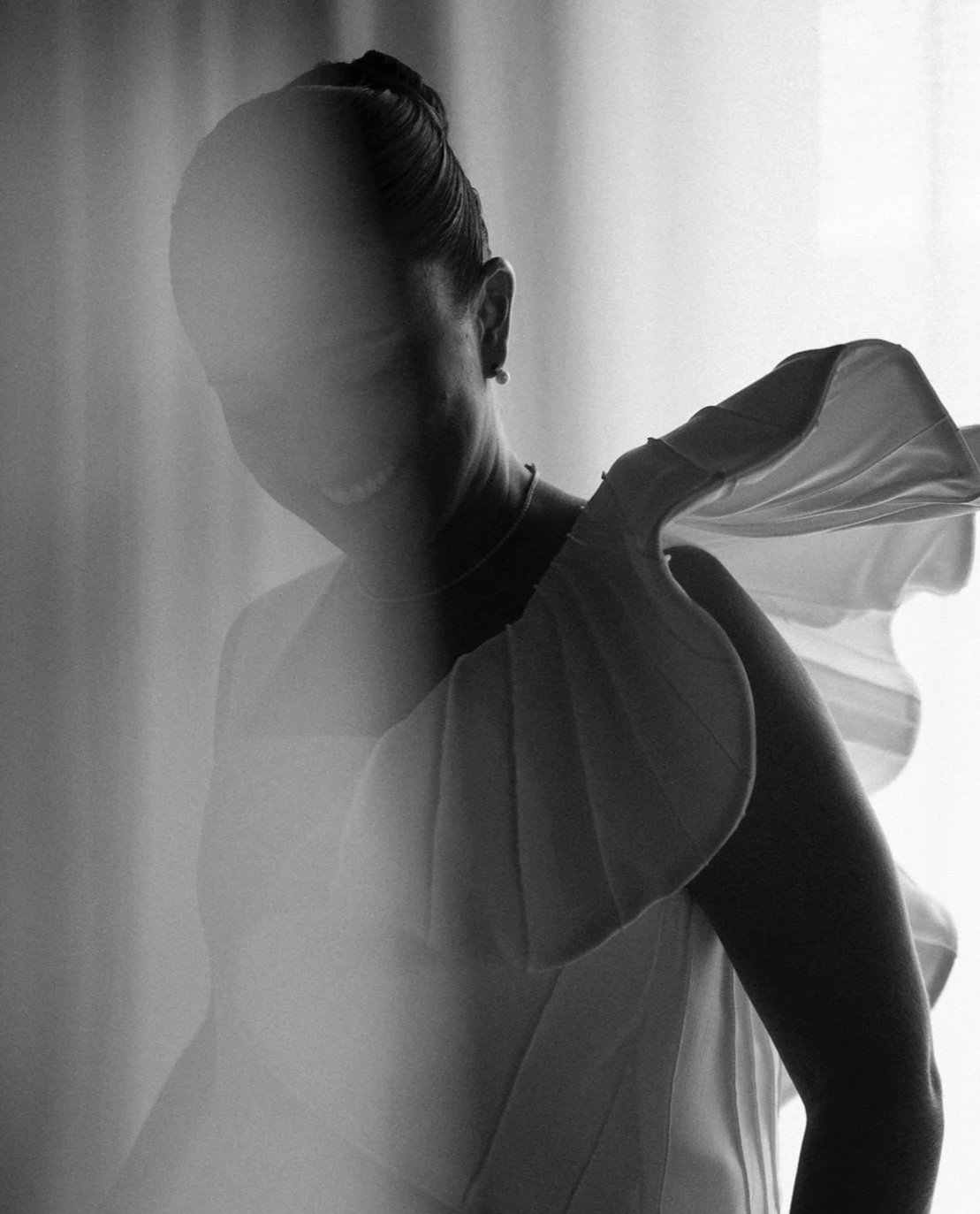Retrato preto e branco de uma mulher com cabelo preso, usando uma saia ou vestido de ombro só, com uma luz suave vindo de uma janela e refletindo na parede ao fundo.
