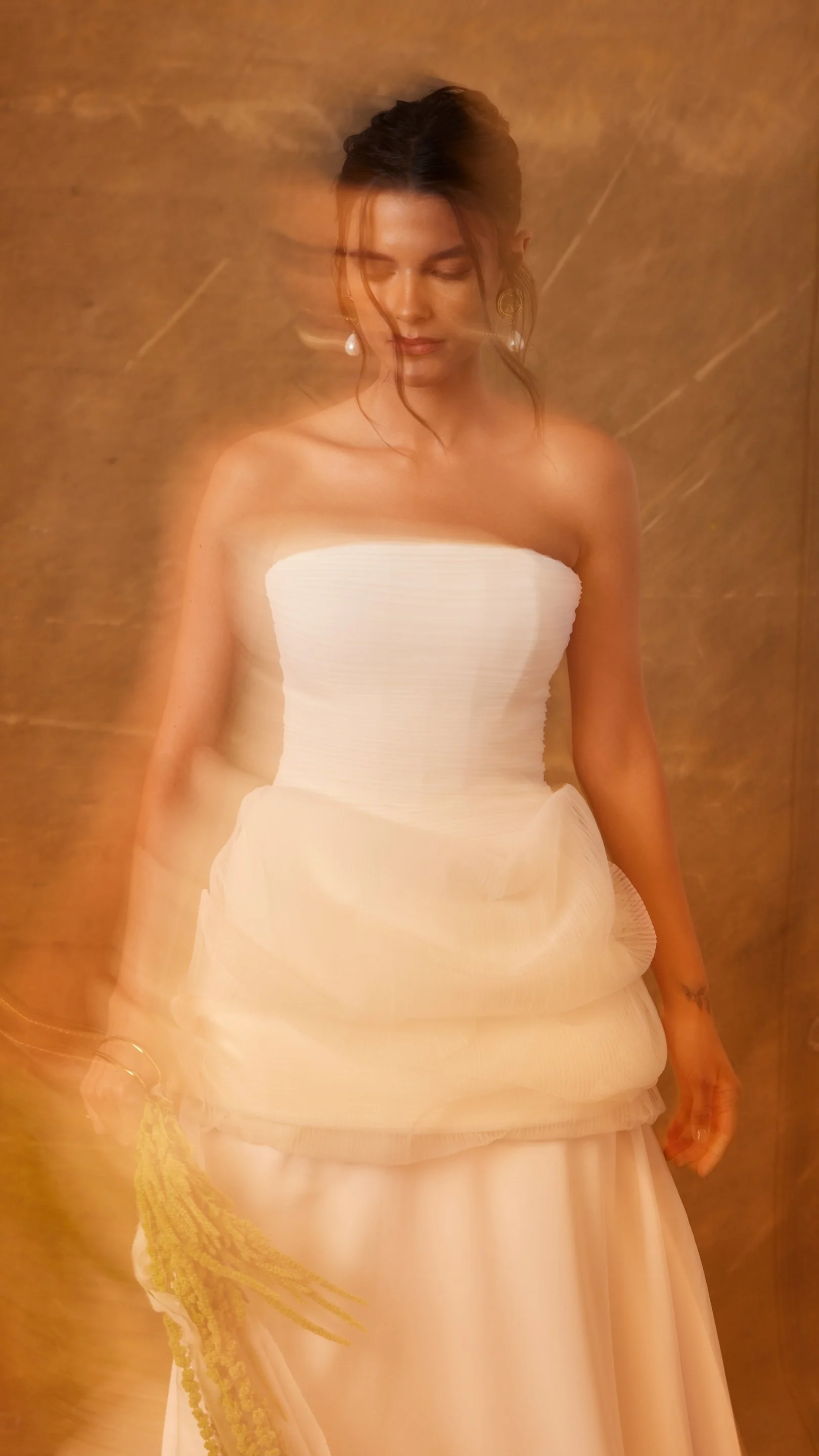 Mulher vestida com vestido branco, com efeito de movimento na foto, segurando um ramos de flores amarelas, com fundo de parede de cor amadeirada.