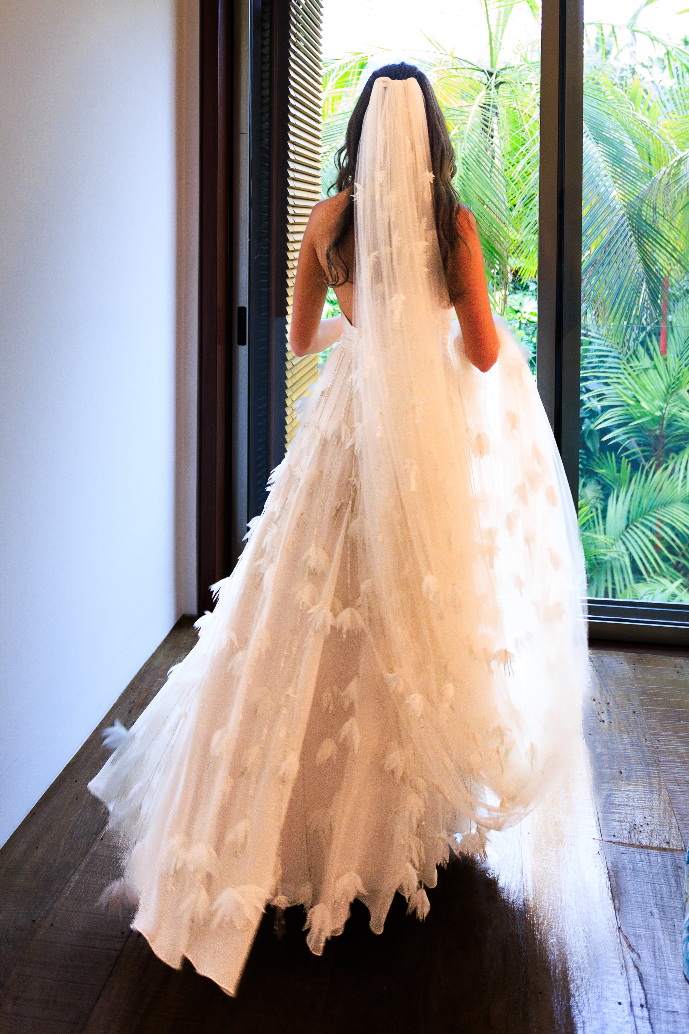 Mulher em vestido de noiva com véu, de costas, olhando para uma janela com vista para plantas verdes.