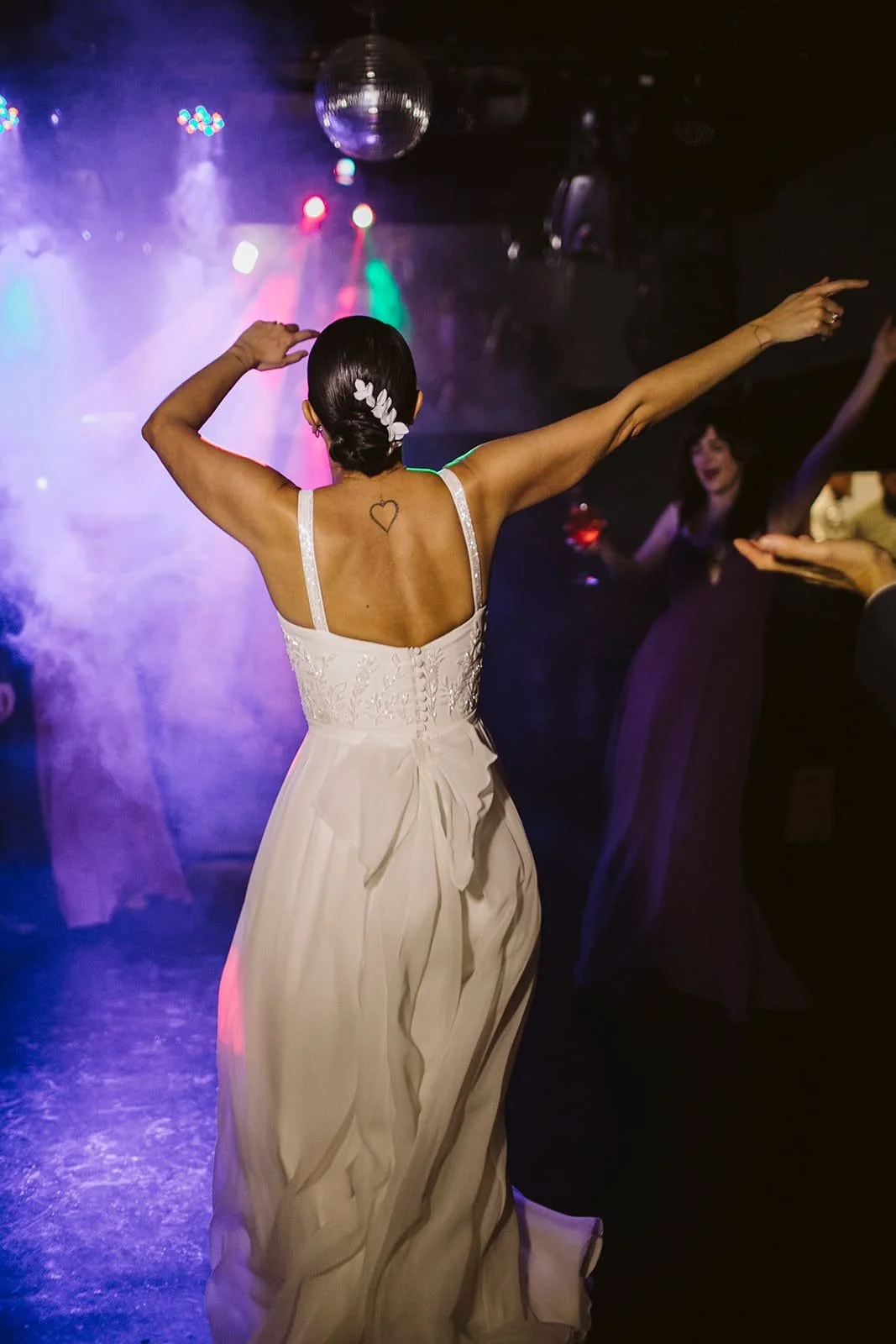 Mulher com vestido branco e cabelo escuro em casamento ou festa dançando a noite num ambiente com luzes coloridas e uma bola de espelho no teto.