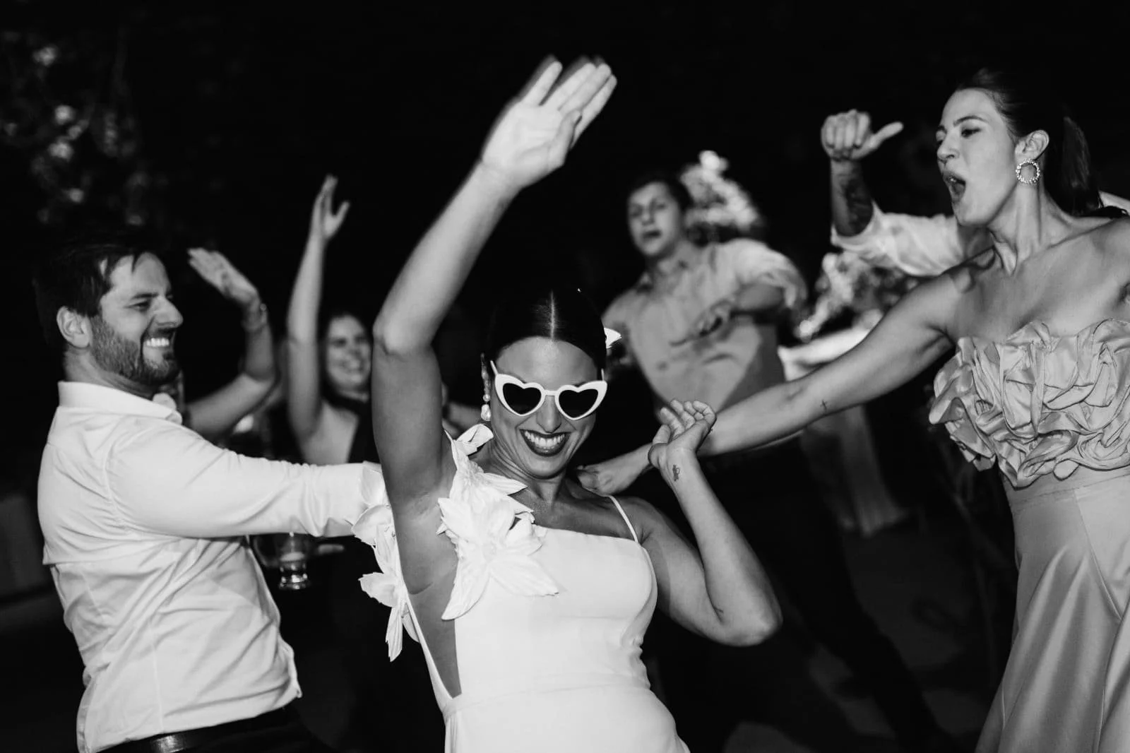 Mulher sorrindo com óculos de armação em forma de coração, dançando com um grupo durante uma festa.