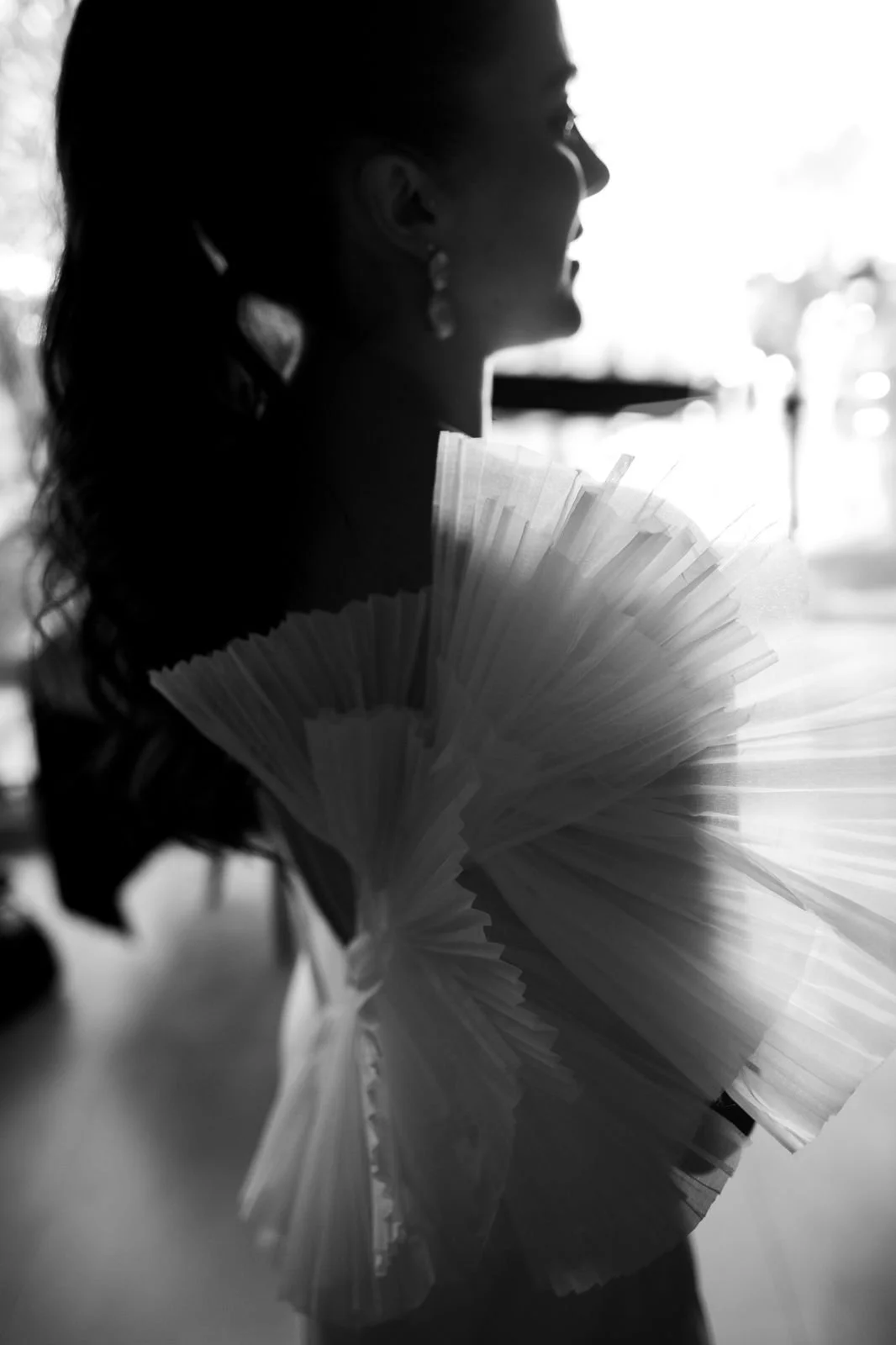 Silueta de uma mulher com cabelo ondulado preso em rabo de cavalo, usando brincos grandes, segurando um leque de papel, em uma imagem em preto e branco, com fundo desfocado.