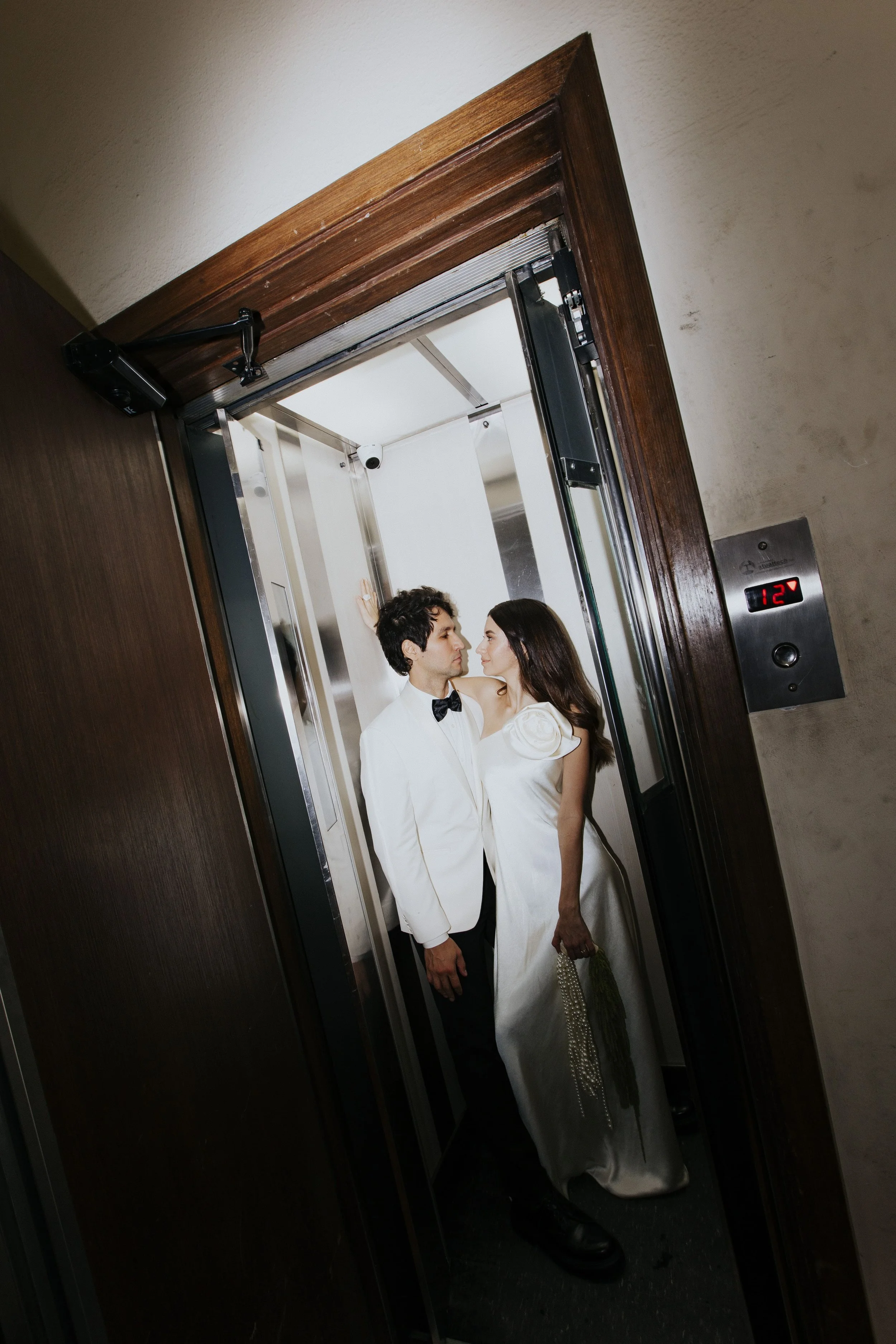 Casal de noivos dentro de um elevador, olhando um para o outro, com roupas de casamento, a mulher segurando um bouquet de flores.