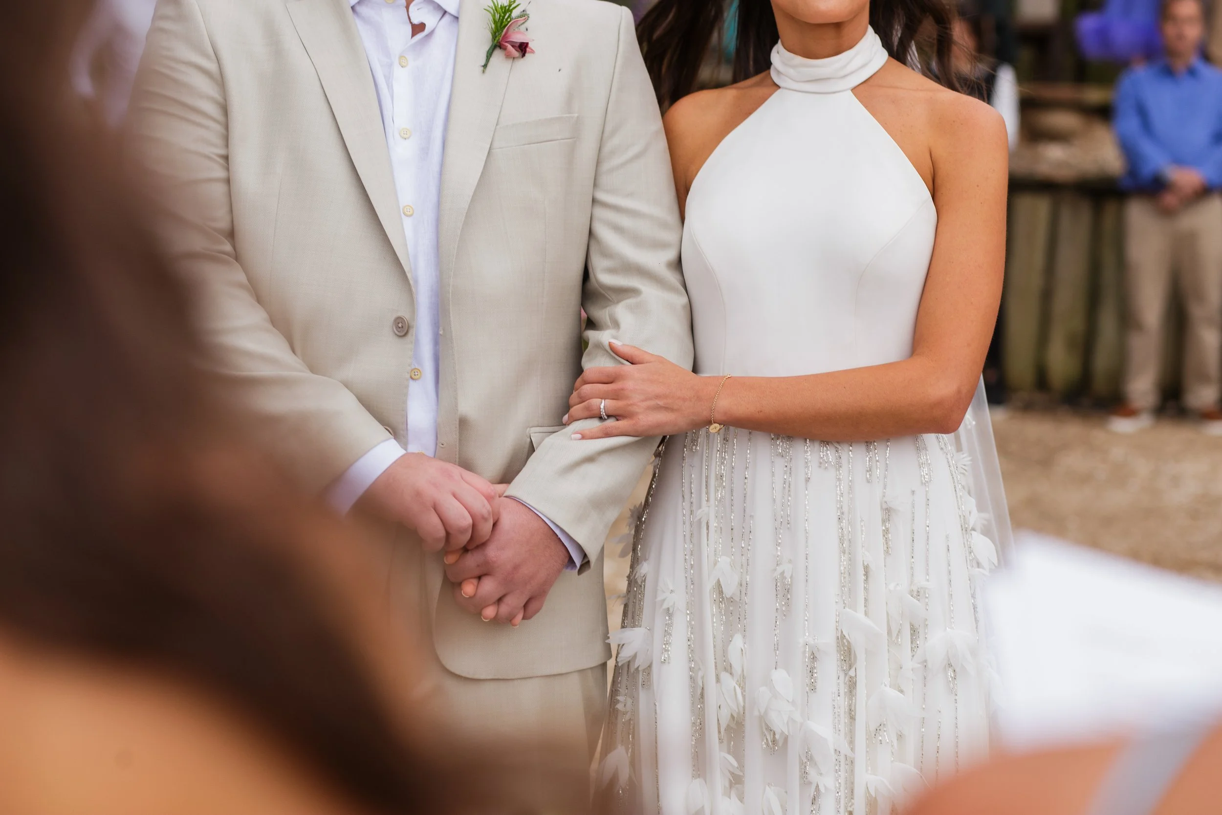 Casal de noivos diante do altar, com o homem vestindo terno bege e a mulher vestindo vestido branco de casamento. Os dois segurando as mãos.
