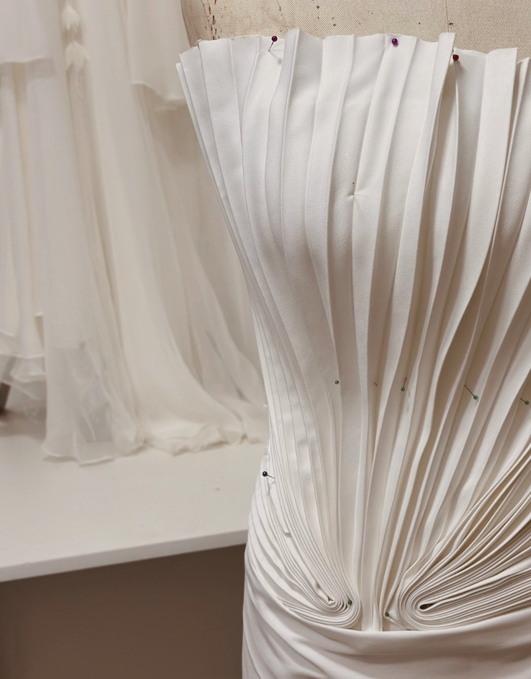 Detalhes de uma saia ou vestido de saia feito em tecido branco, com pregas e pontos de alfinetes na parte superior e inferior.