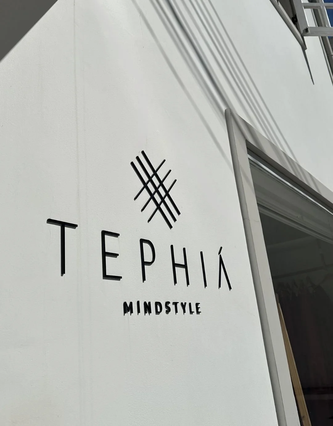 Logo da loja ou marca 'TepHÁ' com a palavra 'Mindstyle' abaixo, em uma parede branca, ao lado de uma janela e com sombras de fios ou cabos na parede.