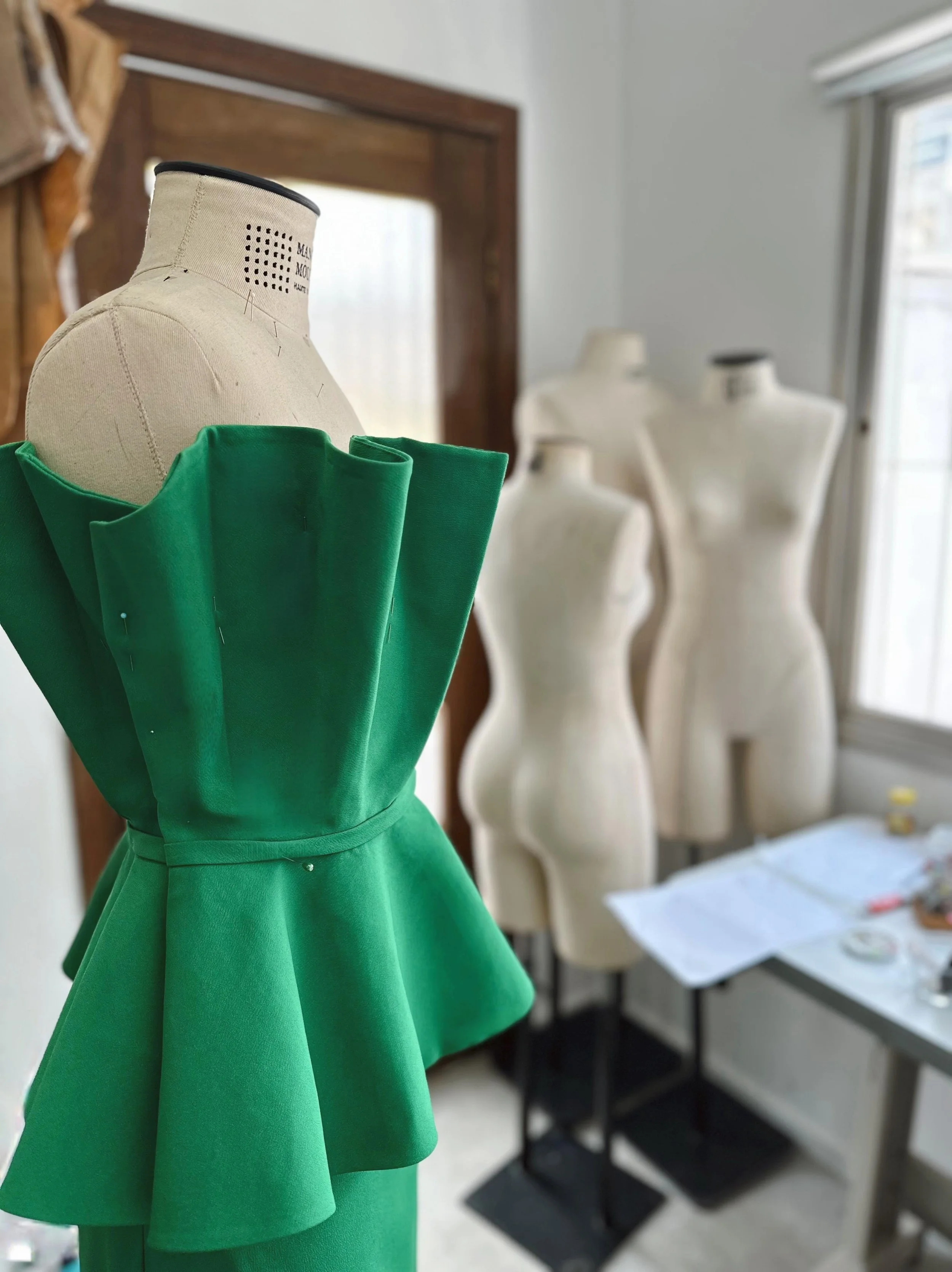Manequim com vestido verde estilo peplum em um ambiente de ateliê de moda, com outros manequins ao fundo e mesa com tecidos e materiais de costura.