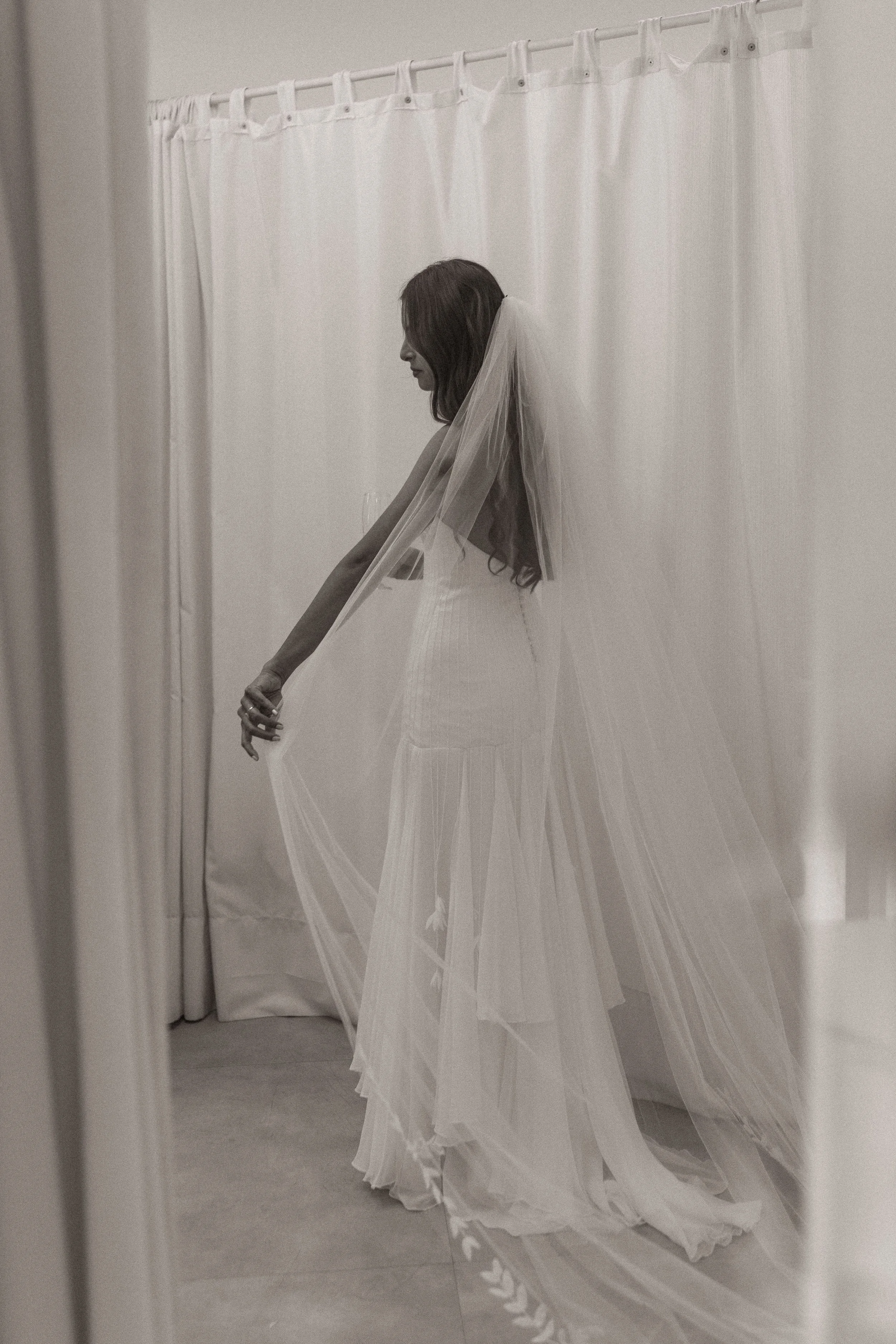 Mulher vestida com vestido de noiva e véu, segurando a renda da saia, de costas, em um ambiente com cortinas brancas.