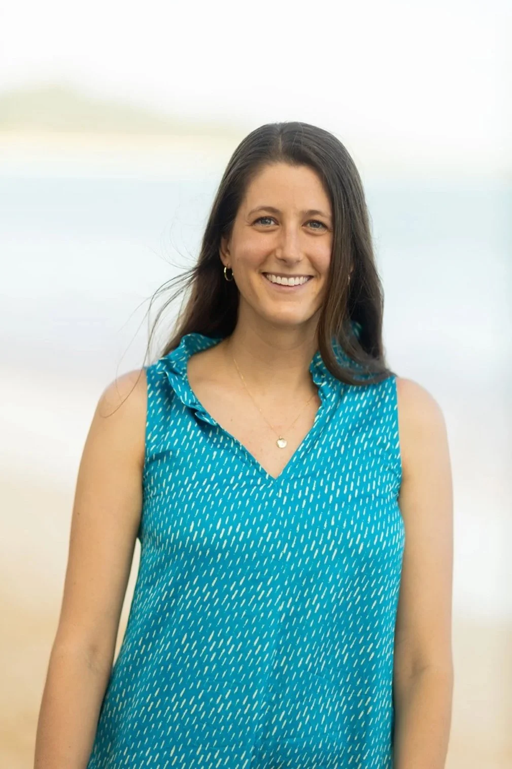 Katey, a woman with long brown hair smiling at the camera, wearing a sleeveless turquoise dress with a small white pattern, standing near on Kailua Beach, Hawai'i, with a blurred background.