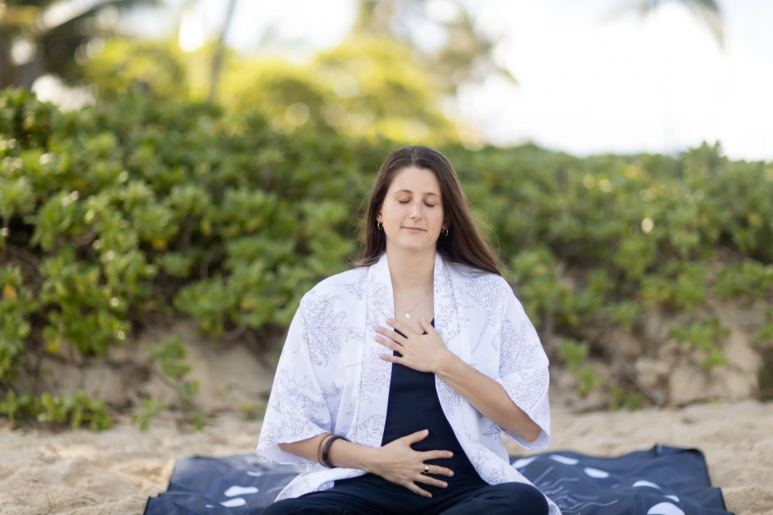 Katey practicing meditation at Kailua Beach, Hawai'i with closed eyes, sitting cross-legged on a mat, wearing a white kimono over a navy jumper