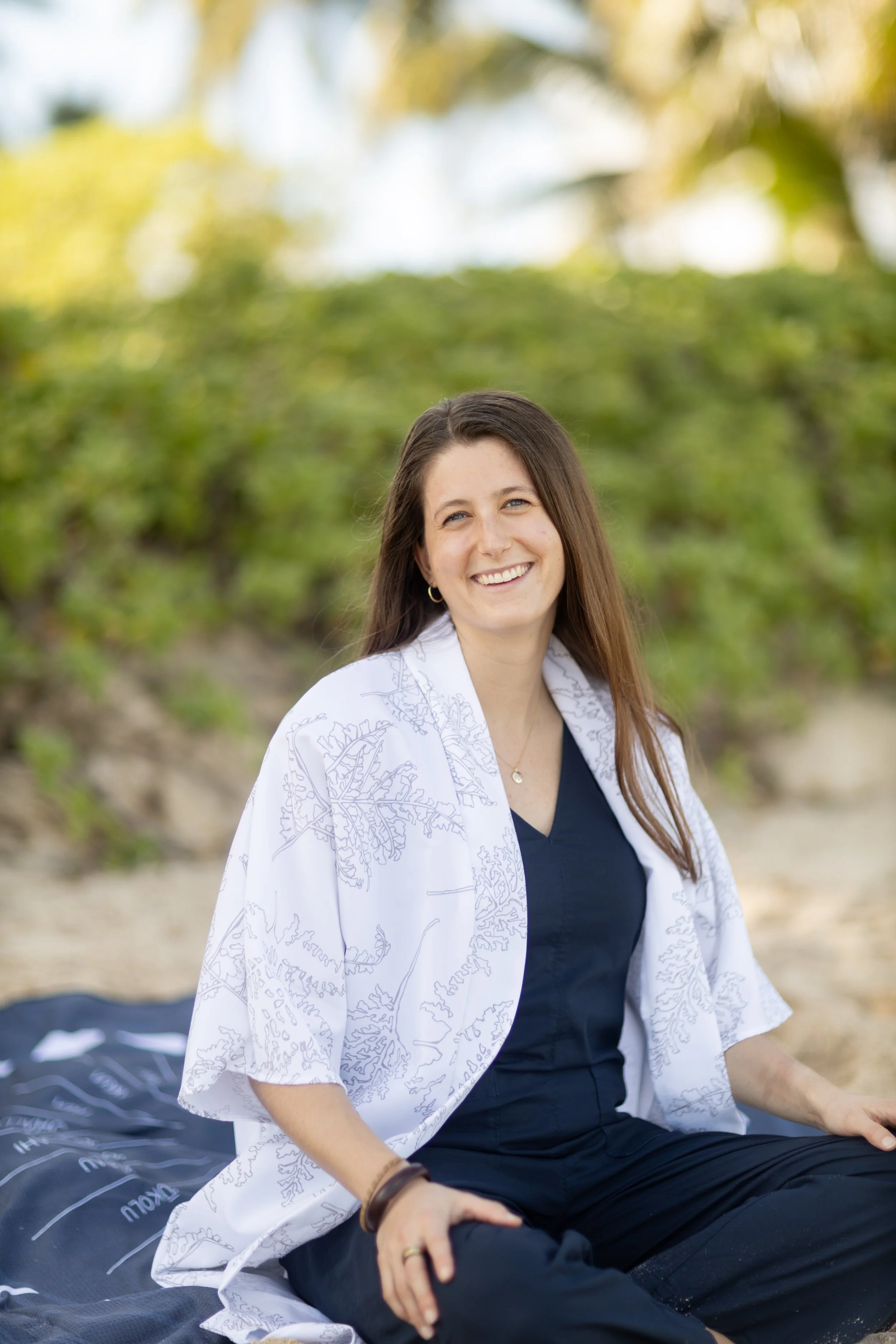 Katey, a woman with long brown hair and a white patterned kimono, sits outdoors at Kailua Beach, Hawai'i with greenery in the background, smiling at the camera.