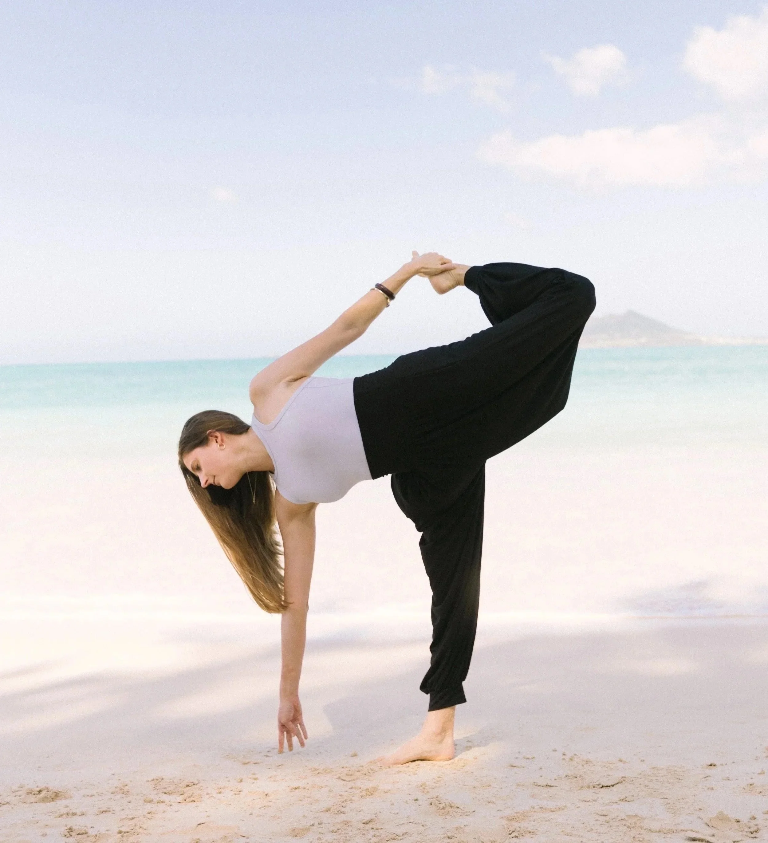 Katey, a woman, practices yoga on a beach, balancing in a one-legged extended hand pose (sugarcane) near the shoreline of Kailua Beach, Hawai'i with ocean and sky in the background.