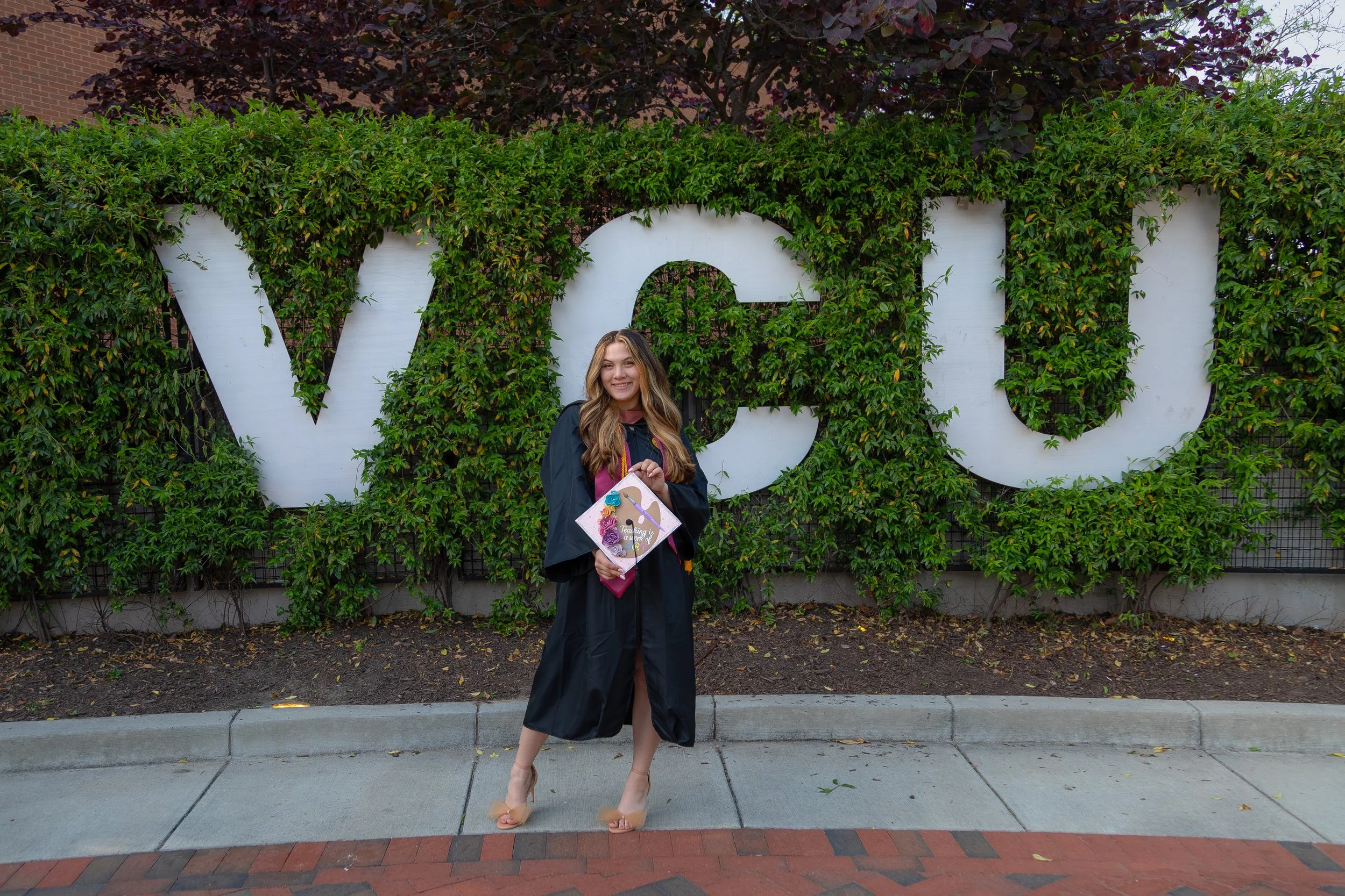 A woman in a graduation gown smiles at the camera while holding her decorated graduation cap.