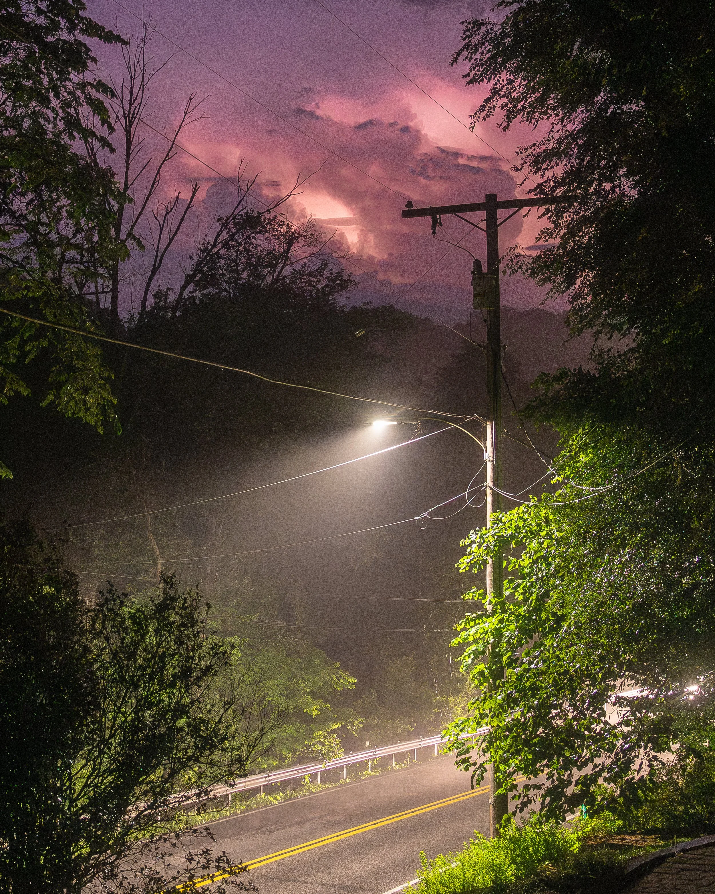 A streetlight illuminates a road wet with a summer storm. The sky is pink with lightning.