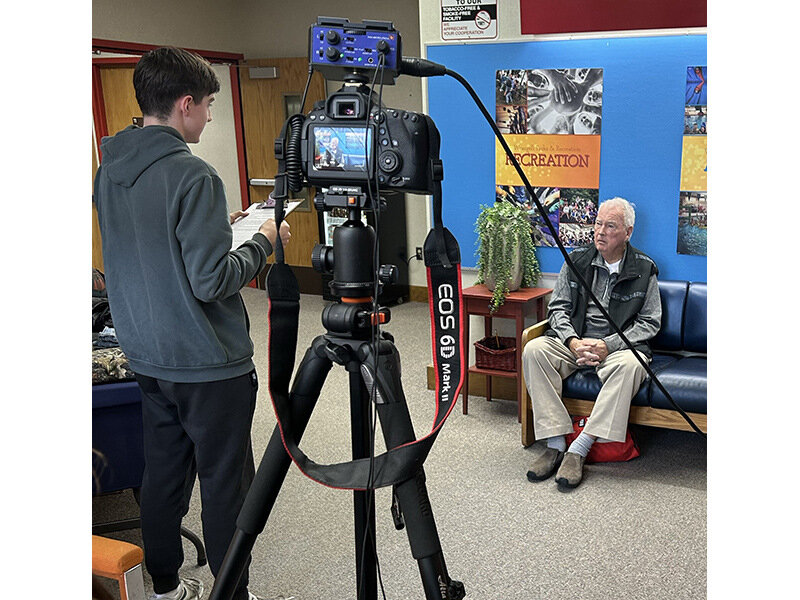 A young man is recording an interview with an older man using a professional camera setup in a room with colorful artwork and a blue wall.