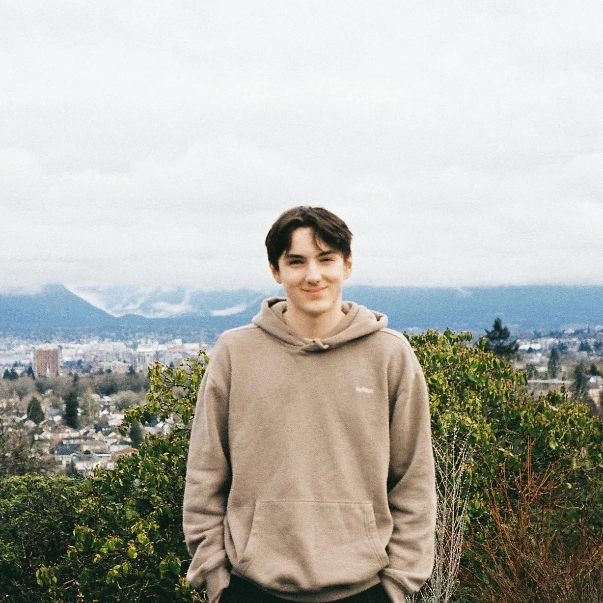 Young man with short dark hair smiling outdoors, wearing a beige hoodie, standing in front of a cityscape with mountains and cloudy sky in the background.