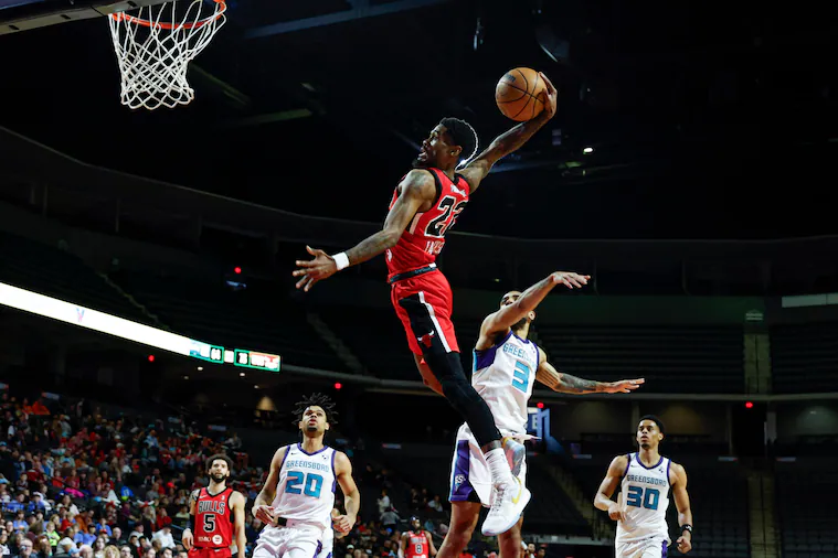 Basketball player in red uniform jumping to dunk the ball during a game, with opponents and spectators in the background.