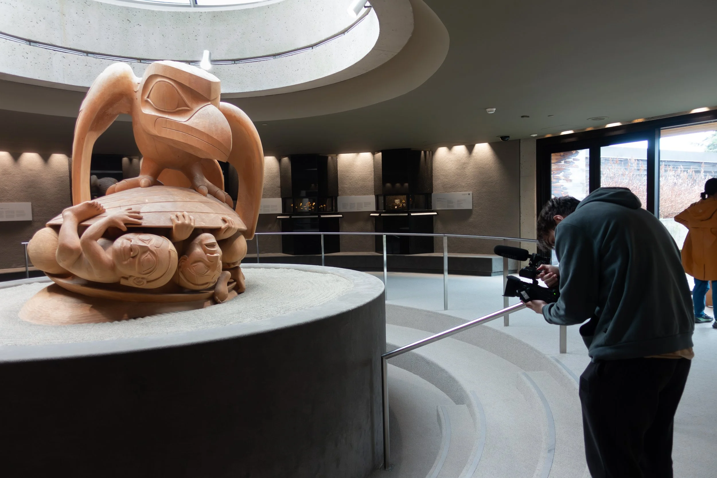A man filming a wooden sculpture inside a modern museum with large windows and spotlights, with other visitors in the background.