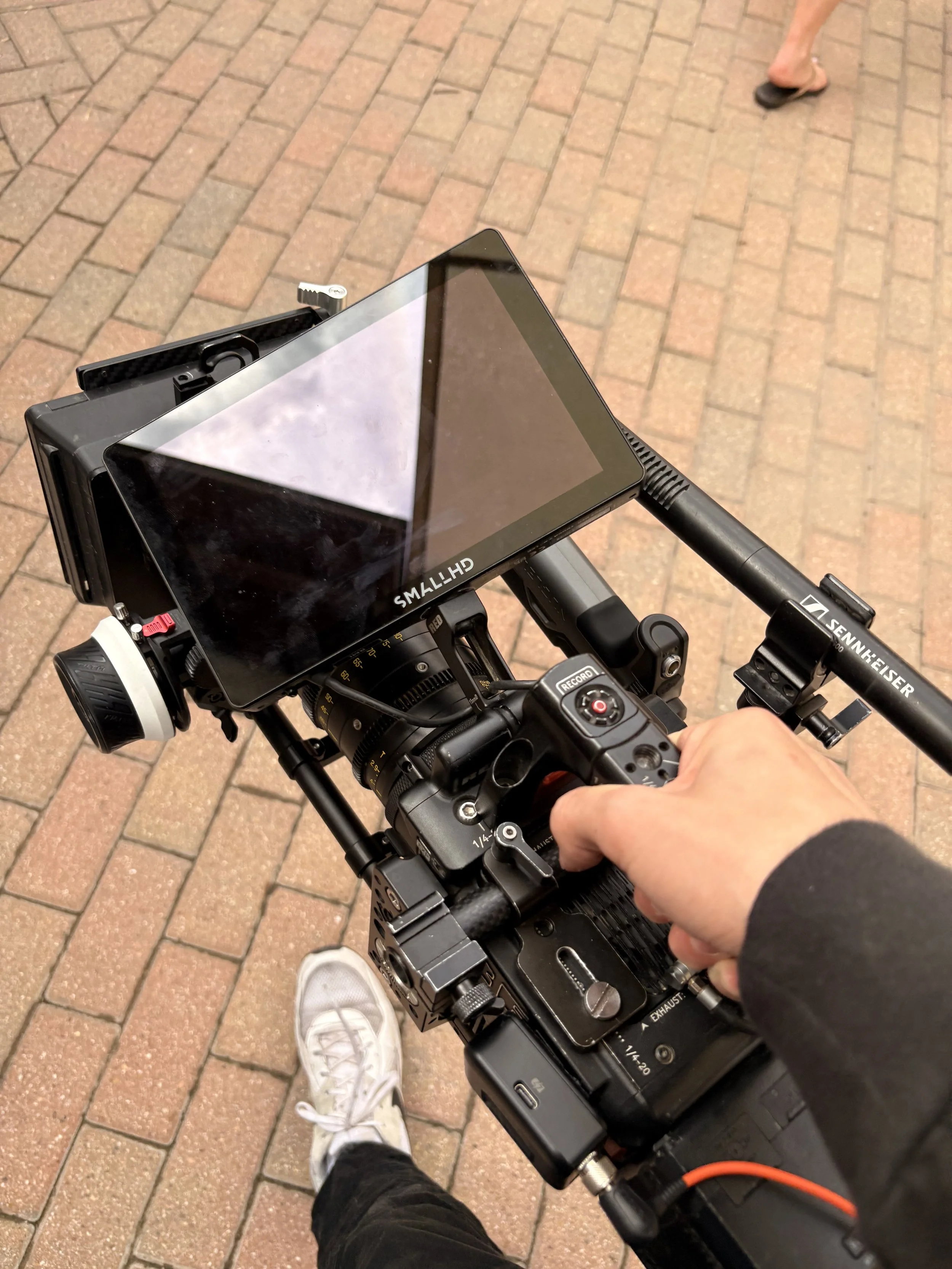 Close-up of a film camera on a dolly with a monitor attached,  on a brick sidewalk, with a person wearing white sneakers and black pants.
